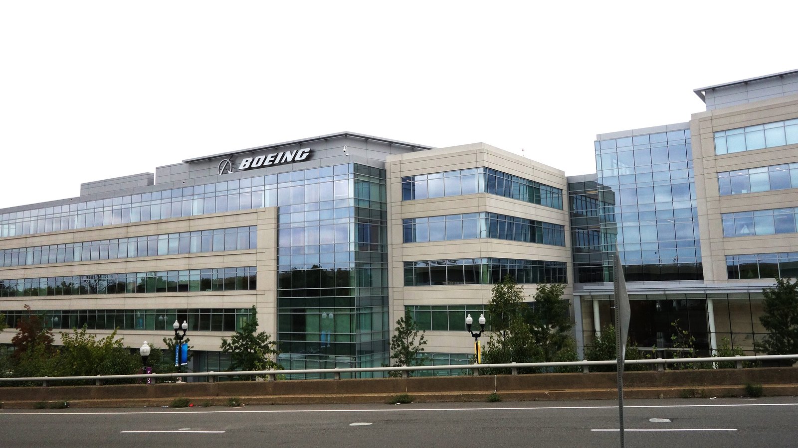 A low green-glass and pale stone office building seen from across a road behind a guard rail, with the Boeing name and its winged emblem mounted on the roofline under a flat overcast sky.