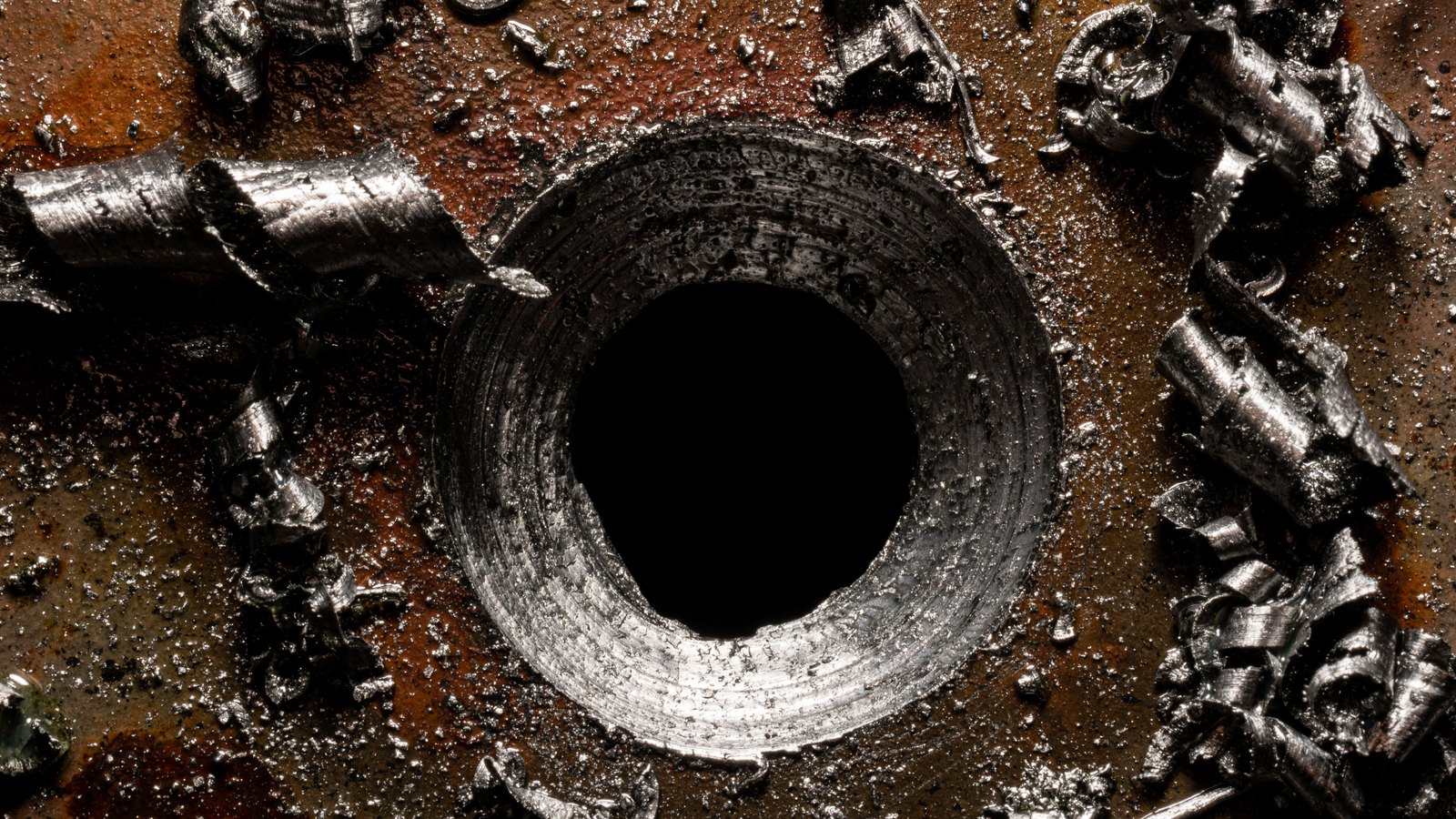 A close view of a freshly drilled hole in a sheet of metal, ringed by bright curled swarf against a dark oiled surface.