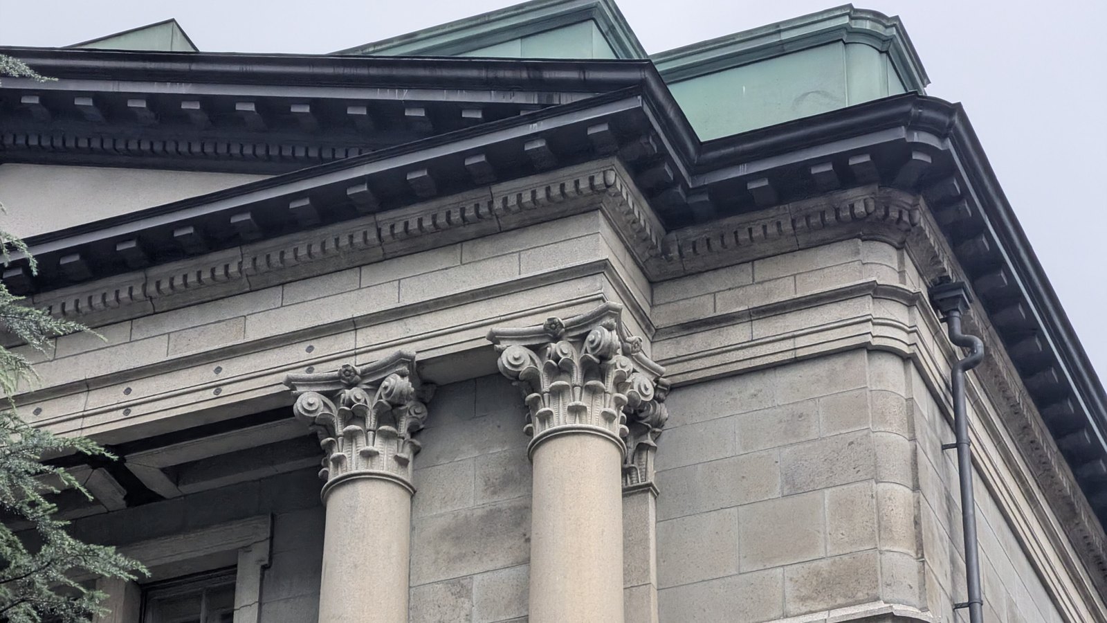 The upper corner of the Bank of Japan head office in Tokyo against a flat grey sky: two Corinthian capitals carrying a heavy black dentilled cornice, coursed granite blocks running back along the return wall, the pale green copper roof edge above, a cast iron downpipe at the right and a cedar branch at the left.