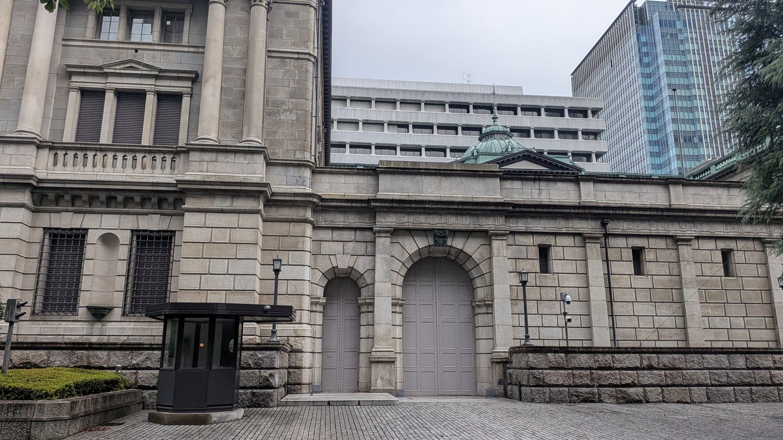 The stone flank of the Bank of Japan head office in Tokyo under flat grey cloud, with two closed arched service gates set into the rusticated wall, an empty black glass guard box on the paving in front, and the building's green copper dome and modern office towers rising behind.