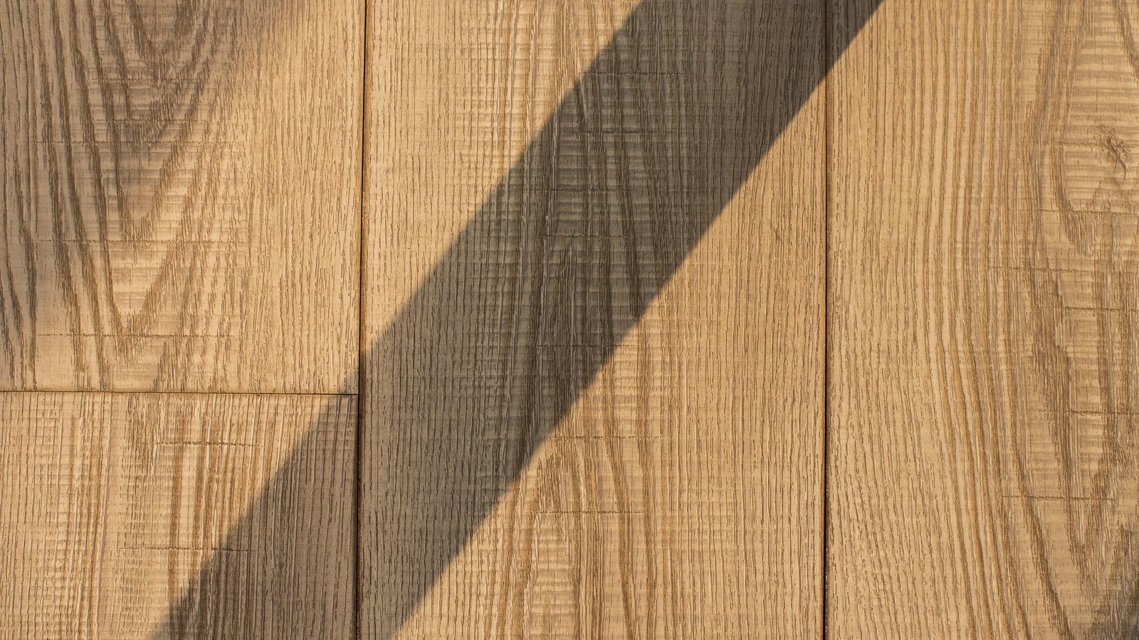 Pale oak floorboards photographed from directly above, with a broad diagonal band of shadow falling across the grain from the lower left to the upper right.