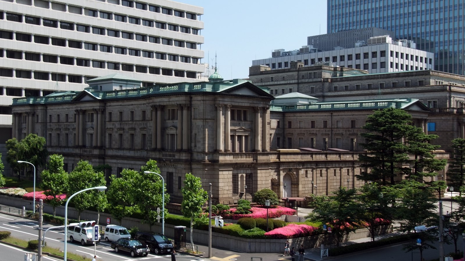 The Bank of Japan head office in Nihonbashi, Tokyo, photographed from above street level on a clear day: a long stone neoclassical building with a green copper roof and a columned portico, banked azaleas and street trees along its wall, and taller modern office blocks rising behind it.