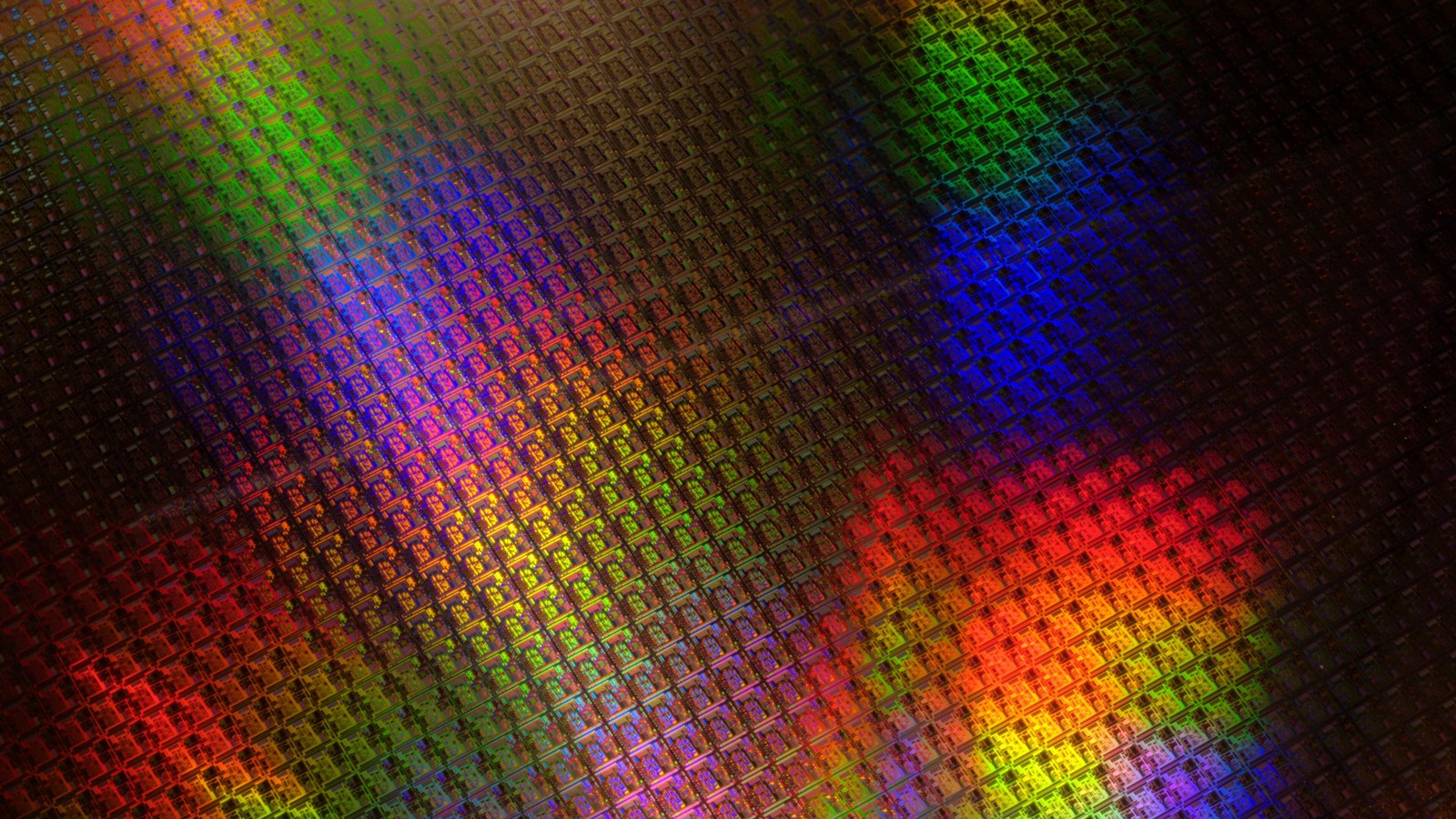 A close macro of a finished silicon wafer filling the frame with rows of identical rectangular dies, the internal circuit blocks of each one visible. Diffraction off the polished surface throws the grid into diagonal bands of red, orange, yellow, green, blue and violet against darker areas between them. No text, lettering, logo, wafer edge, hand or tool is in the picture.