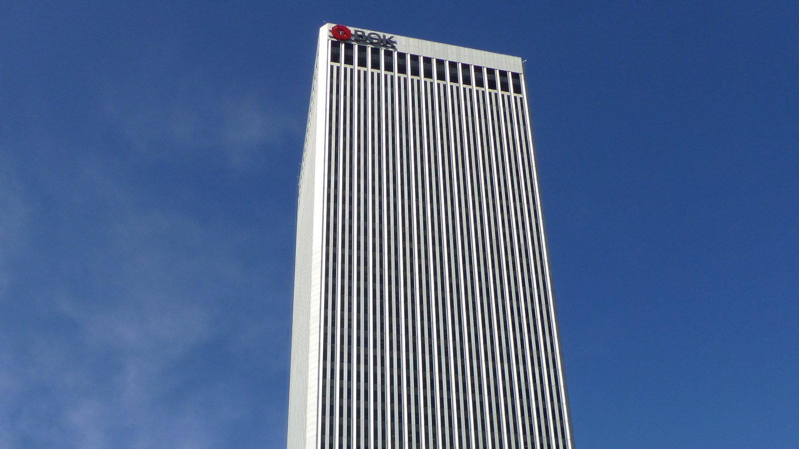 The upper floors of the white BOK Tower in Tulsa, its vertical ribs rising against a deep blue sky, with the BOK name and red logo at the top.