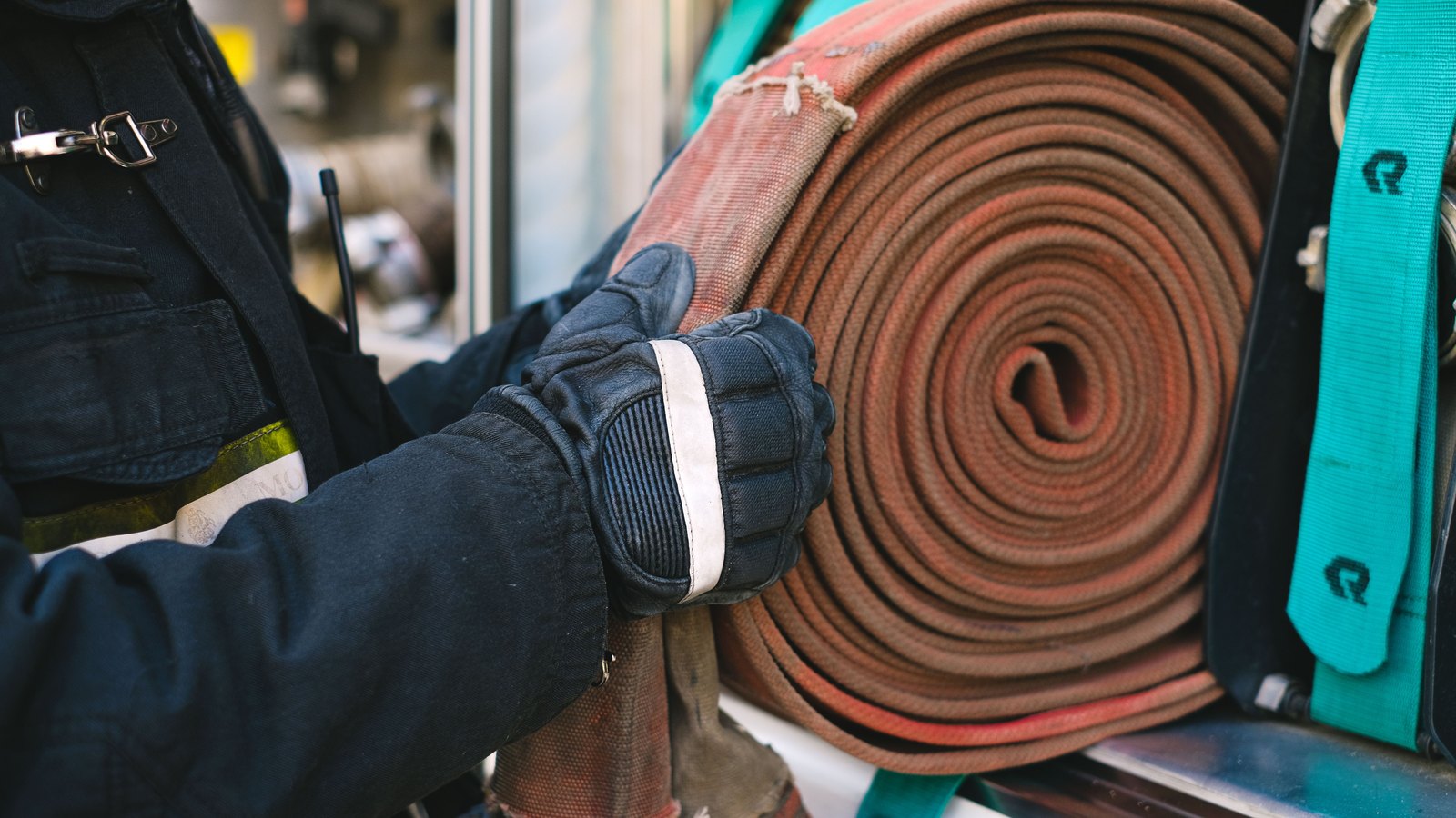 A close view of a firefighter's gloved hands guiding a thick rolled canvas fire hose into a compartment on the side of a fire apparatus. The firefighter wears dark turnout gear with a reflective stripe at the cuff and a radio antenna at the chest; no face is visible. A teal securing strap hangs beside the hose. No agency name, department, unit number, readable licence plate, person's face or identifiable place appears anywhere in the frame. A generic illustration of fire crew equipment, not a photograph of this event.
