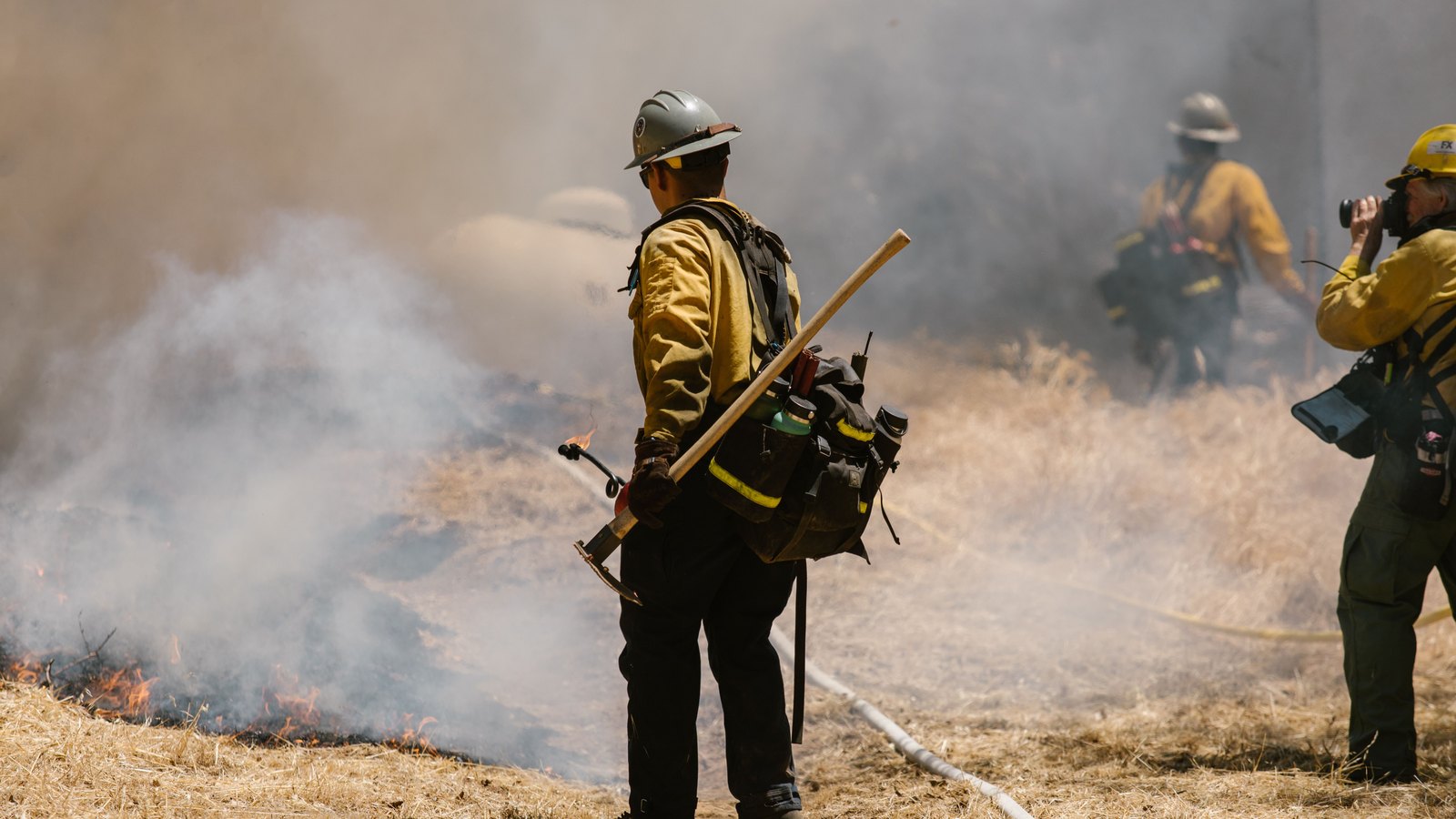 A wildland firefighter in yellow nomex and a plain helmet stands with a hand tool beside a low line of flame creeping through flat dry grass, thick pale smoke drifting across the frame and a charged hose line lying on the ground. Two further crew members are blurred and cropped in the background. No face is identifiable and there is no legible lettering, badge, unit number, plate or place name anywhere in the picture. Stock photograph, not a photograph of this event.