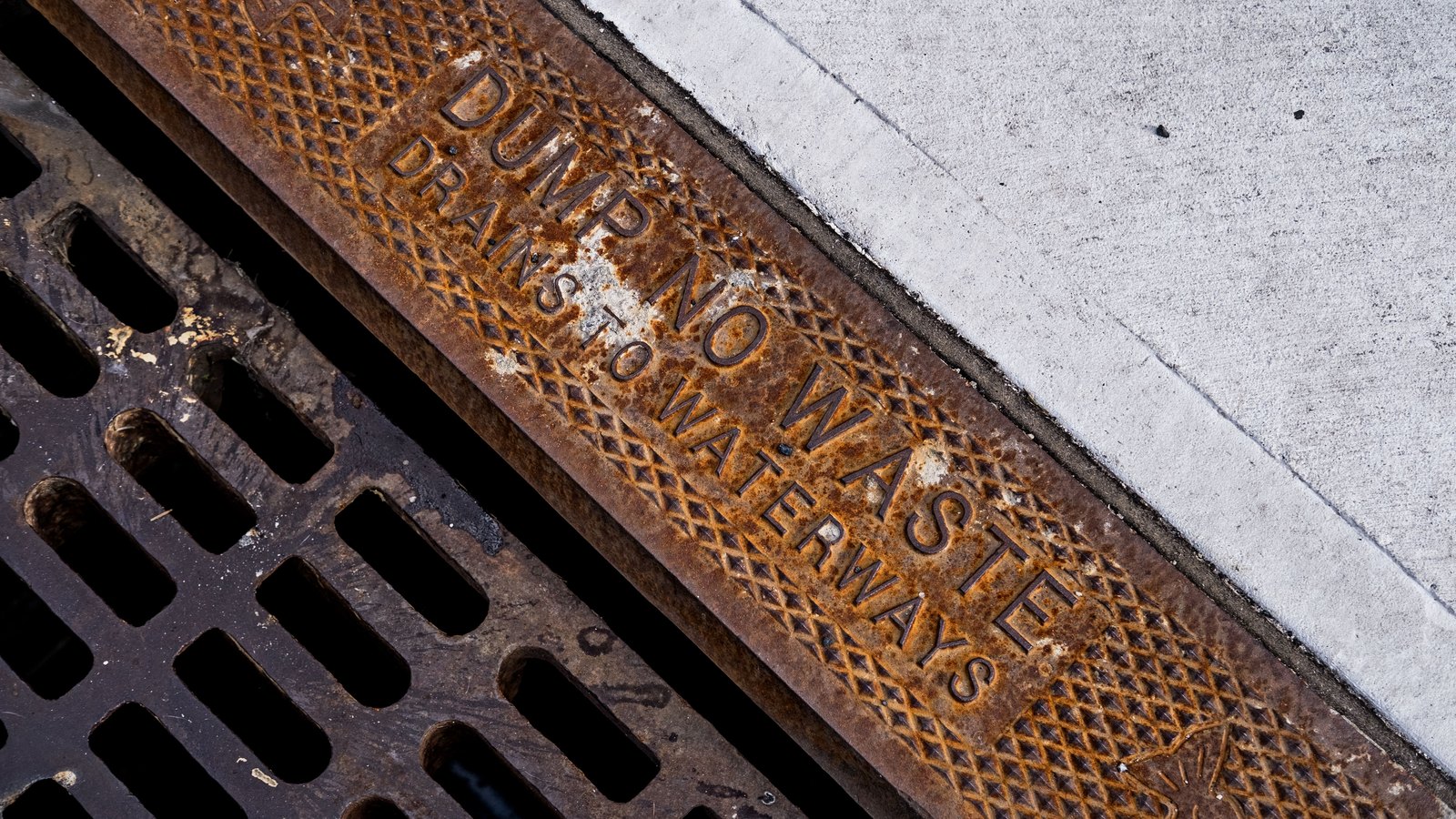 A rusted cast iron storm drain grate at a curb, stamped with the words dump no waste, drains to waterways.