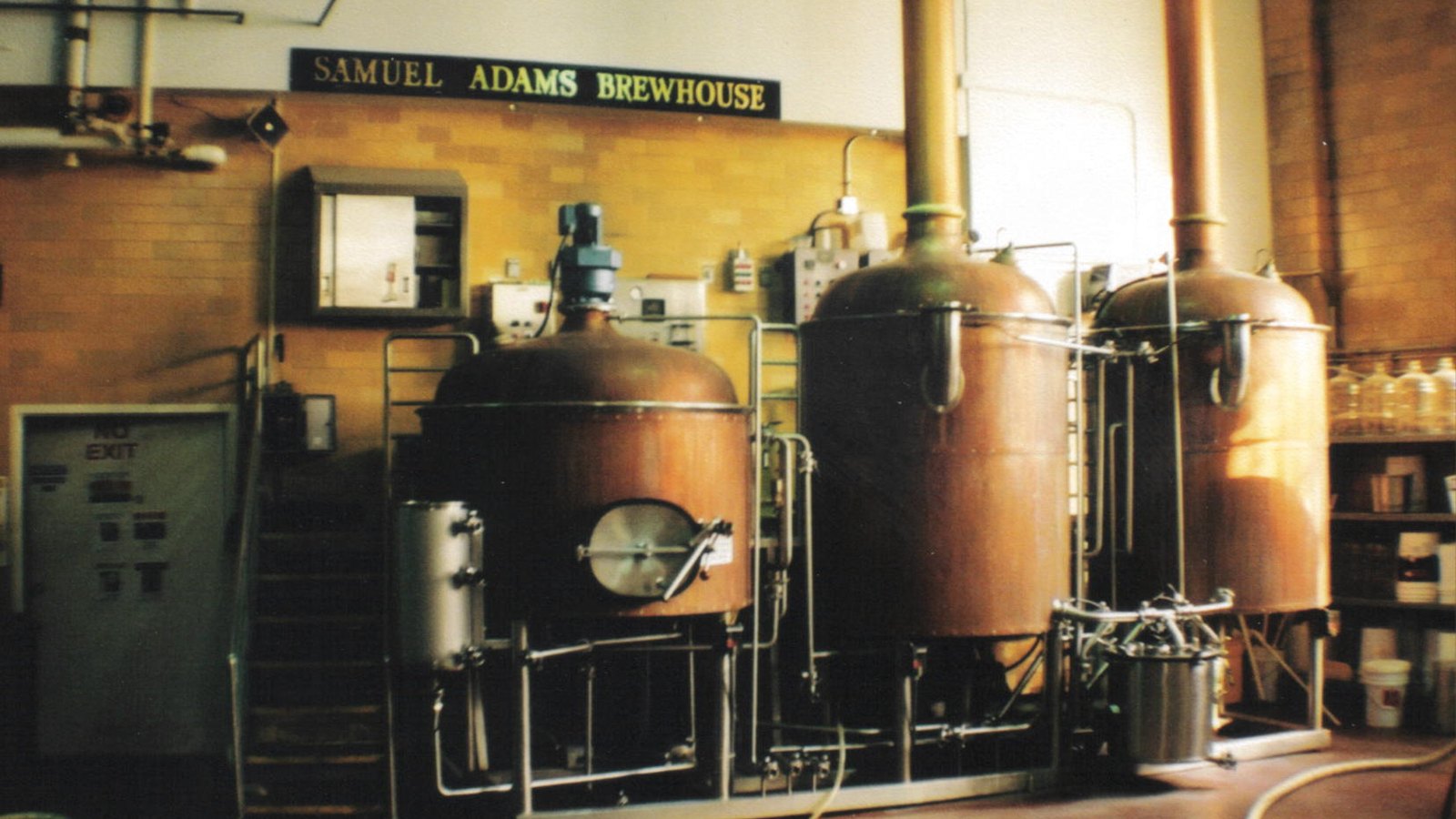 Three copper brew kettles with tall copper flues in the Samuel Adams brewhouse in Boston, under the brewery's own lettered sign on a yellow tiled wall.