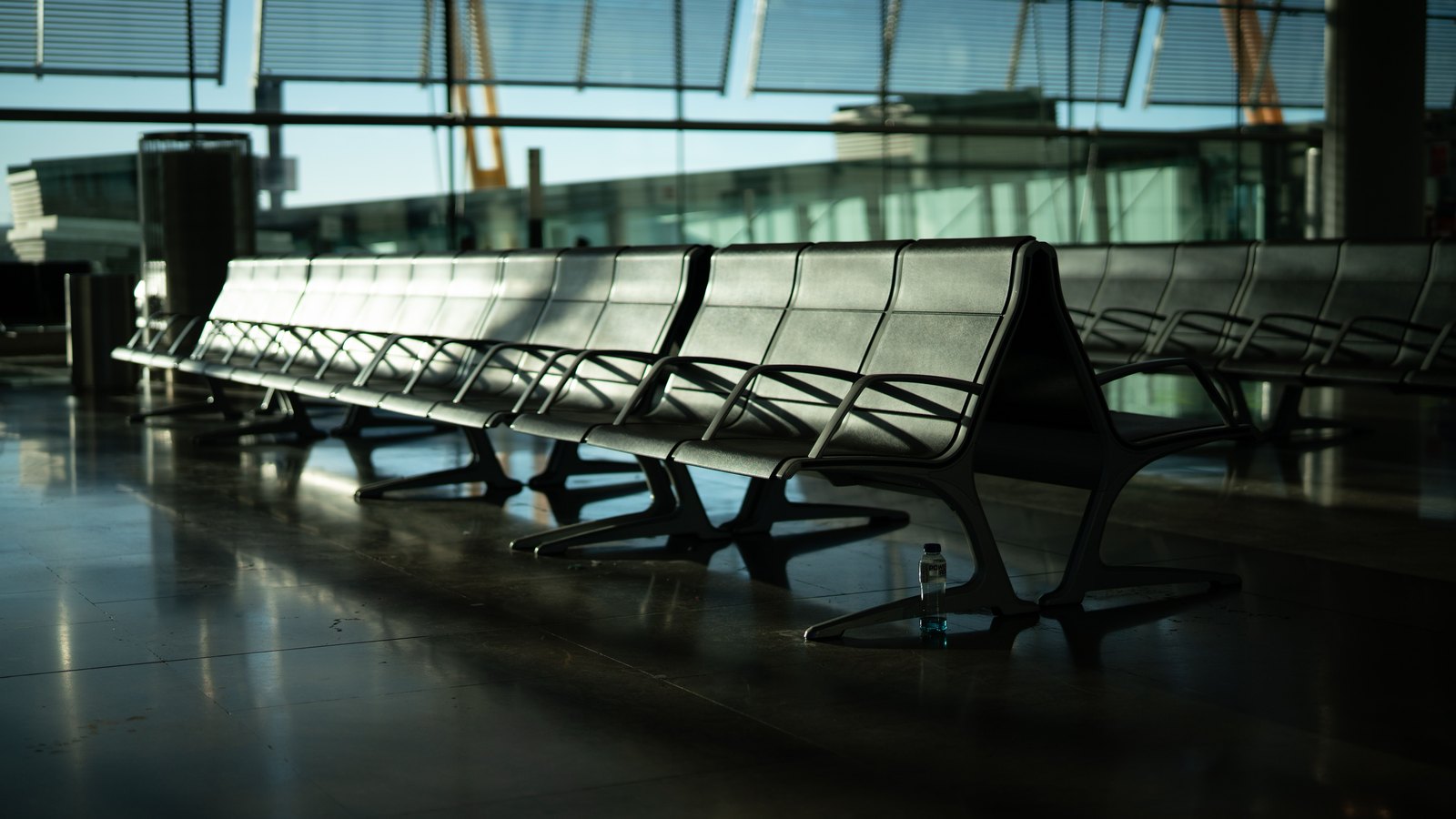 A long empty row of metal bench seating in an airport terminal at dusk, backlit through a tall glass wall, its polished floor reflecting the light. No people, aircraft, airline markings, gate numbers or legible signage appear anywhere in the frame. Stock photograph, not a photograph of this event.