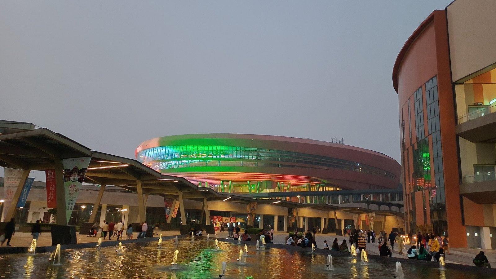 Bharat Mandapam in New Delhi illuminated in green and orange beyond a reflecting pool at dusk.