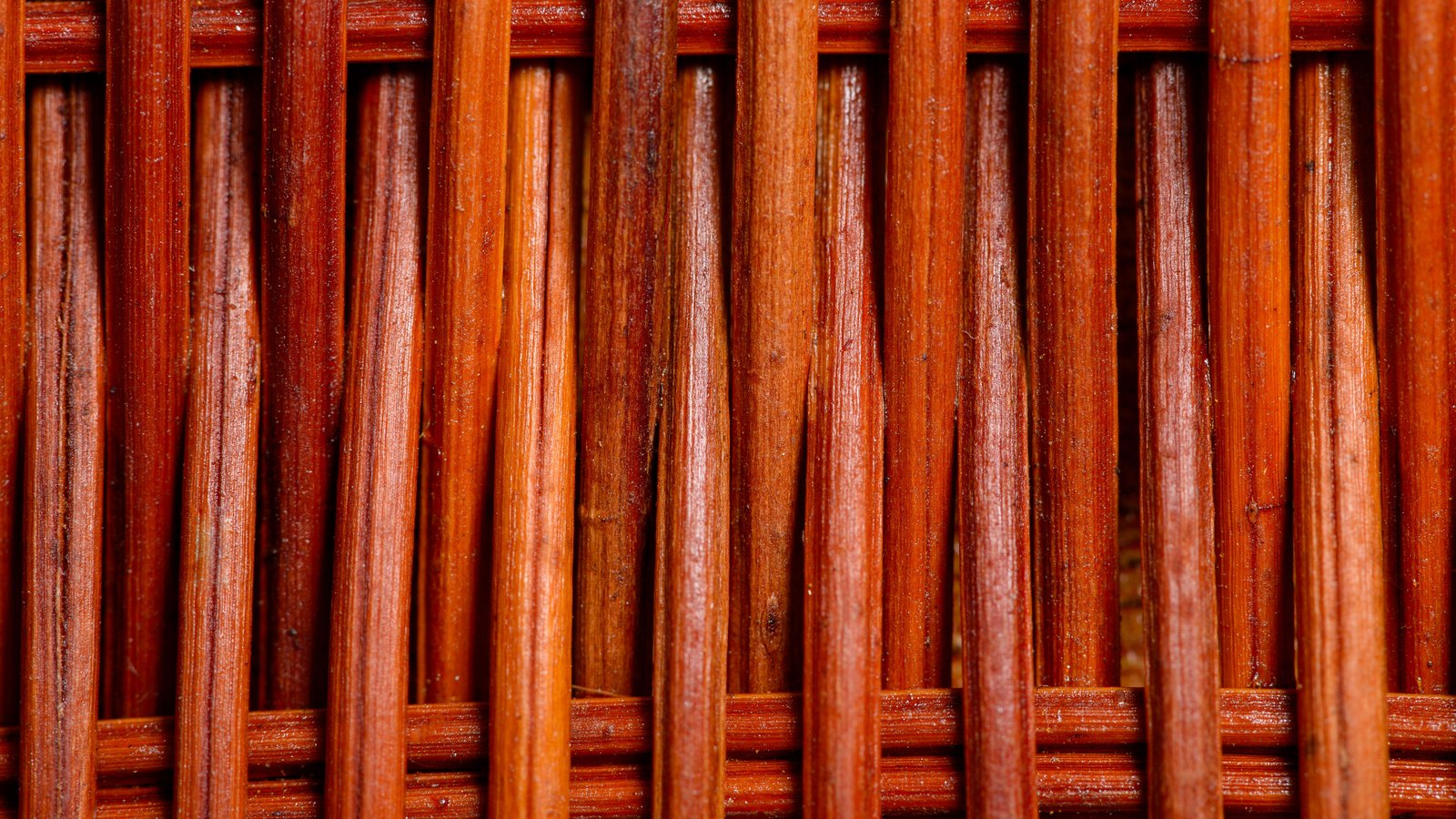 A close photograph of a woven wicker panel, upright rust and amber willow rods pressed tightly side by side and bound top and bottom by horizontal weavers, with narrow dark gaps showing between them. No text, lettering, brand mark, product, person or place is in view.