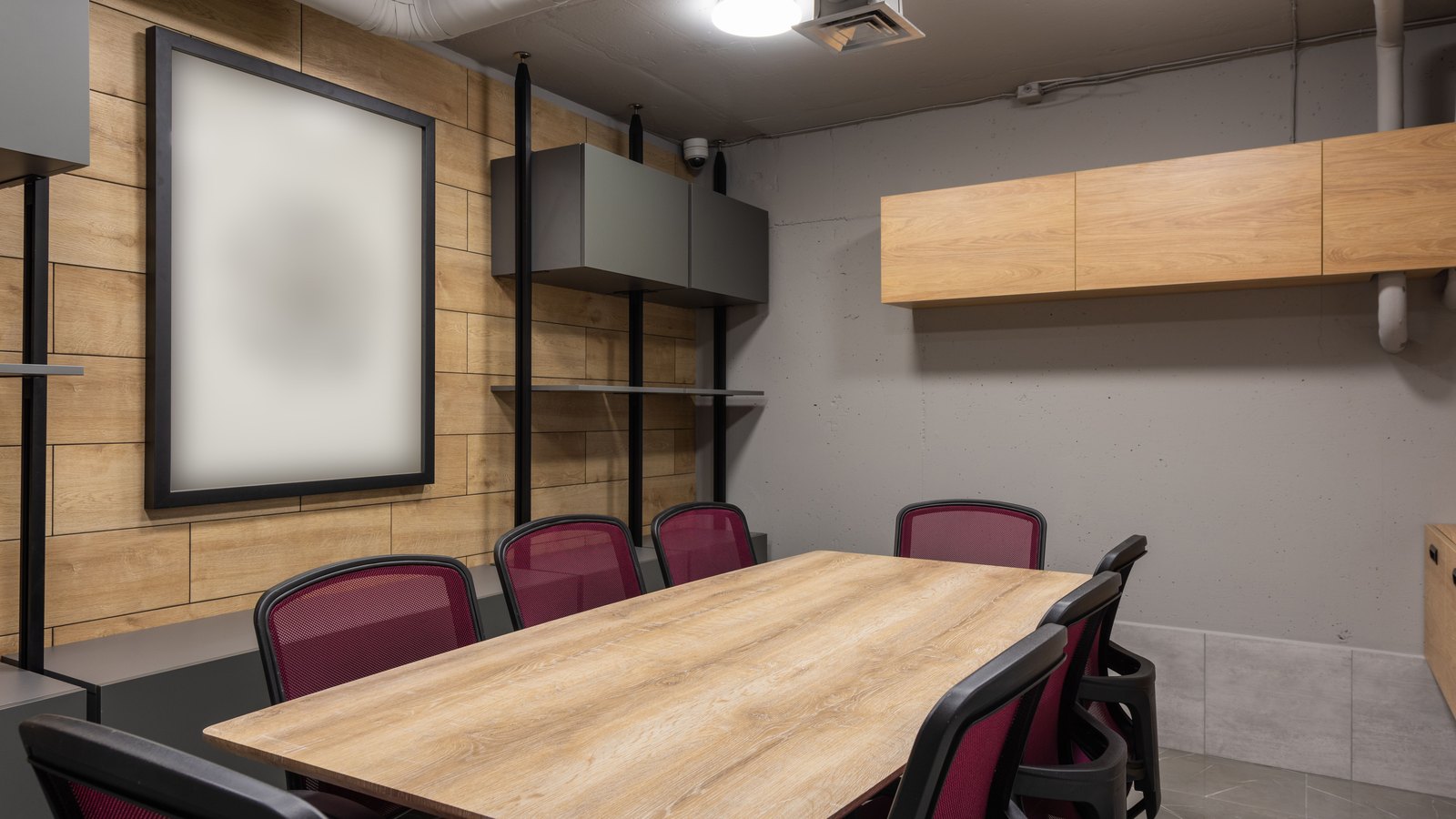 An empty meeting room with a plain wooden table, dark red mesh chairs pushed in around it, a blank framed panel on a timber wall and exposed ducting overhead. No person, text, logo or place is in the frame.