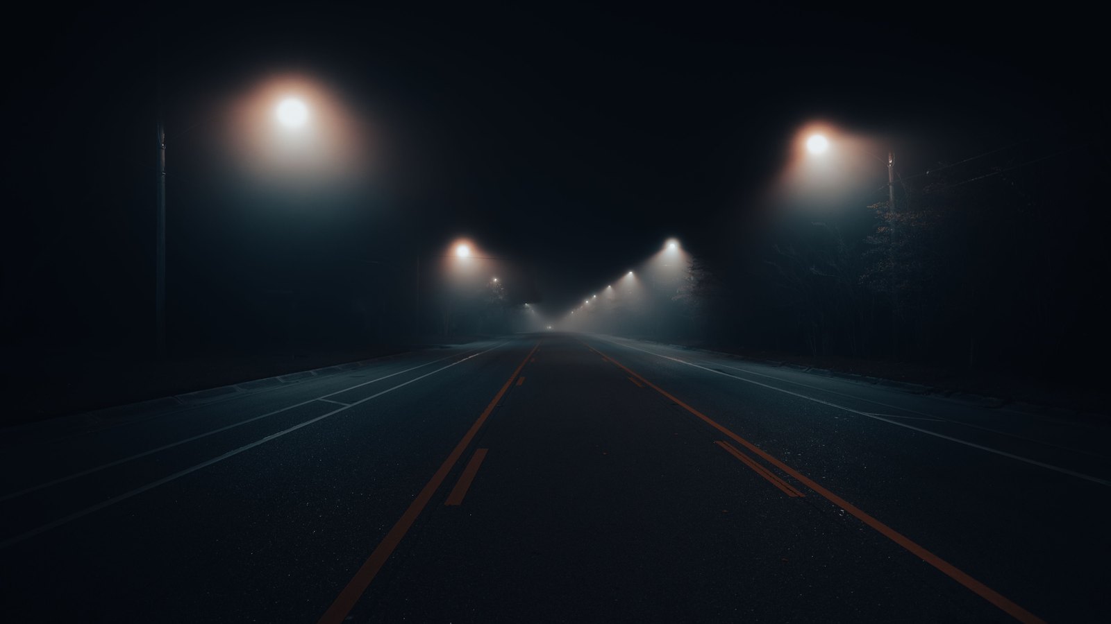 A photograph taken from the centre of an empty asphalt road at night, looking down a straight run of carriageway into mist. Street lights on poles recede toward the horizon on both sides, their beams diffused into halos, and a double yellow centre line runs away from the camera. Bare trees stand in darkness on the right verge. No vehicles, people, signs or lettering are in the frame. A generic illustration, not a photograph of Orange River Loop Road.