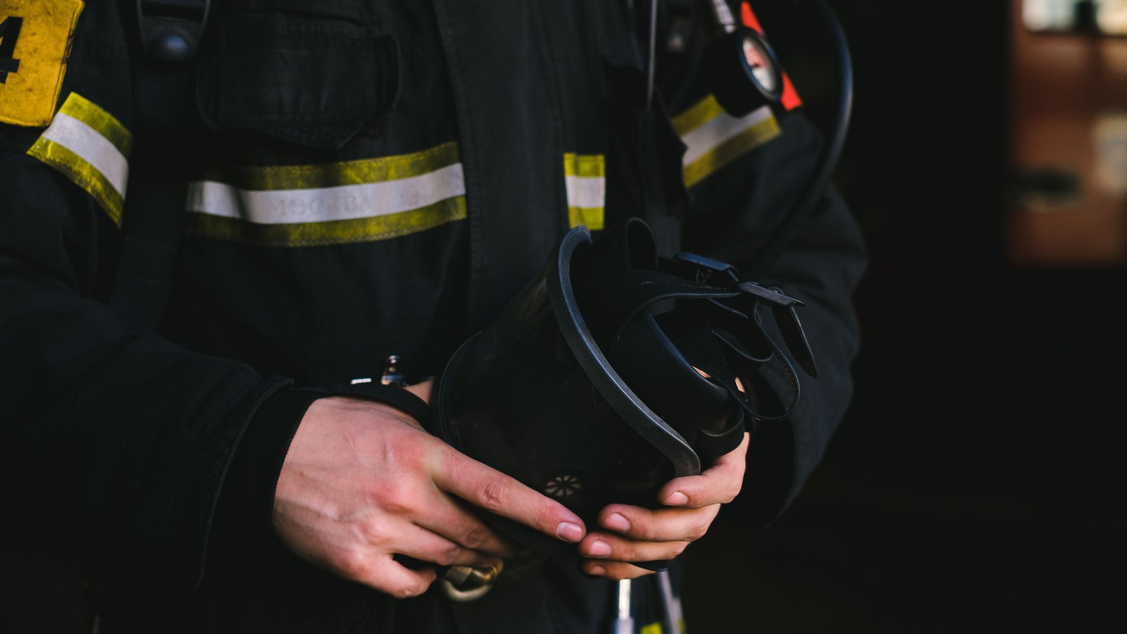 A firefighter in dark turnout gear holds breathing equipment, with the face outside the frame and no agency name visible, not a photograph of this event.