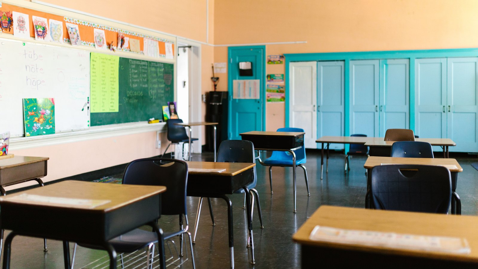 An empty American classroom with rows of tablet-arm desks and moulded chairs, a chalkboard and whiteboard along one wall and pale blue cupboard doors at the back. No child, teacher or other person is in the frame. Handwriting on the boards is out of focus and carries no school name, address, logo or identifying mark of any kind. A generic illustration of a school classroom, not a photograph of either of the centers in this story and not a photograph of this event.