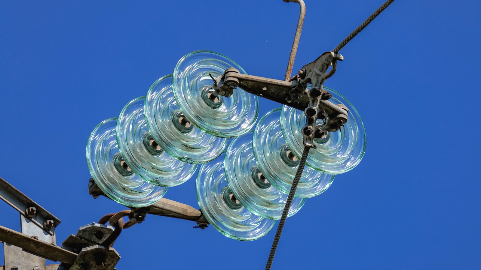 Two strings of toughened glass cap-and-pin insulators, seven discs each, hanging from a weathered steel crossarm against a flat deep blue sky, with a stranded conductor running down through the clamps at the right. No lettering, plate, logo or landmark is visible.