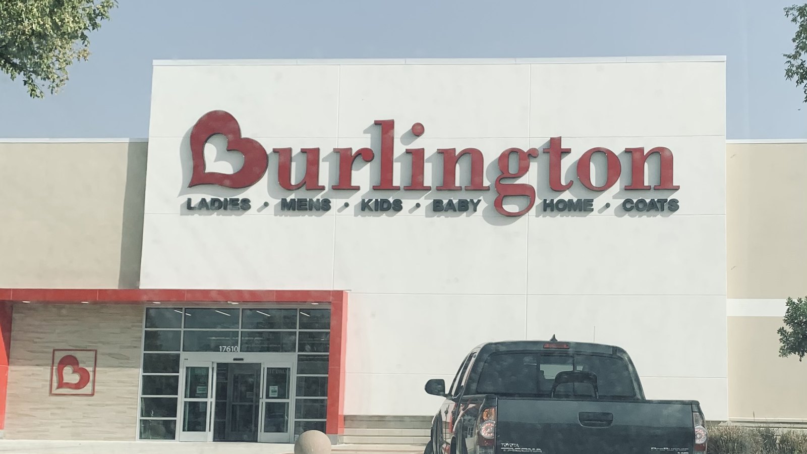 The front of a Burlington store in San Antonio, Texas, photographed from the car park: a plain white and beige box building with the red Burlington wordmark and heart logo across the upper wall, the line LADIES, MENS, KIDS, BABY, HOME, COATS beneath it in dark grey, glass entrance doors under a red canopy, and a dark pickup truck parked in the foreground.