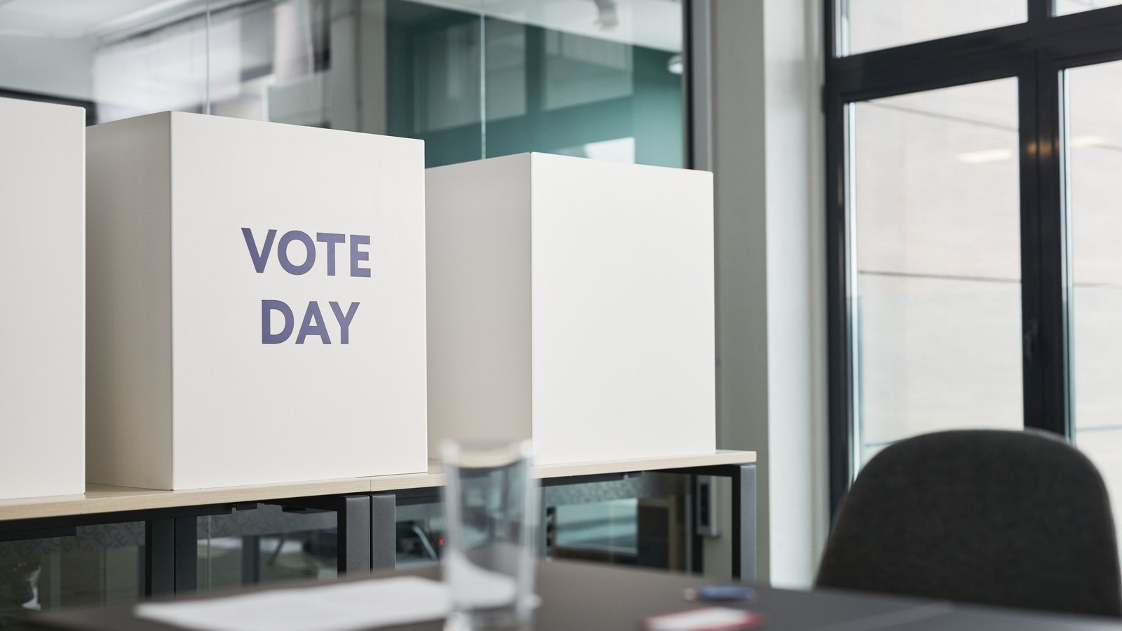 Two white cardboard voting booth partitions standing on a light wooden counter in a bright office with glass partitions and a tall window behind. The nearer screen is printed with the words VOTE DAY in grey capitals. No people are in the frame and nothing identifies a location. It is a stock photograph of polling booths, not a photograph of any Collier or Lee County polling place.