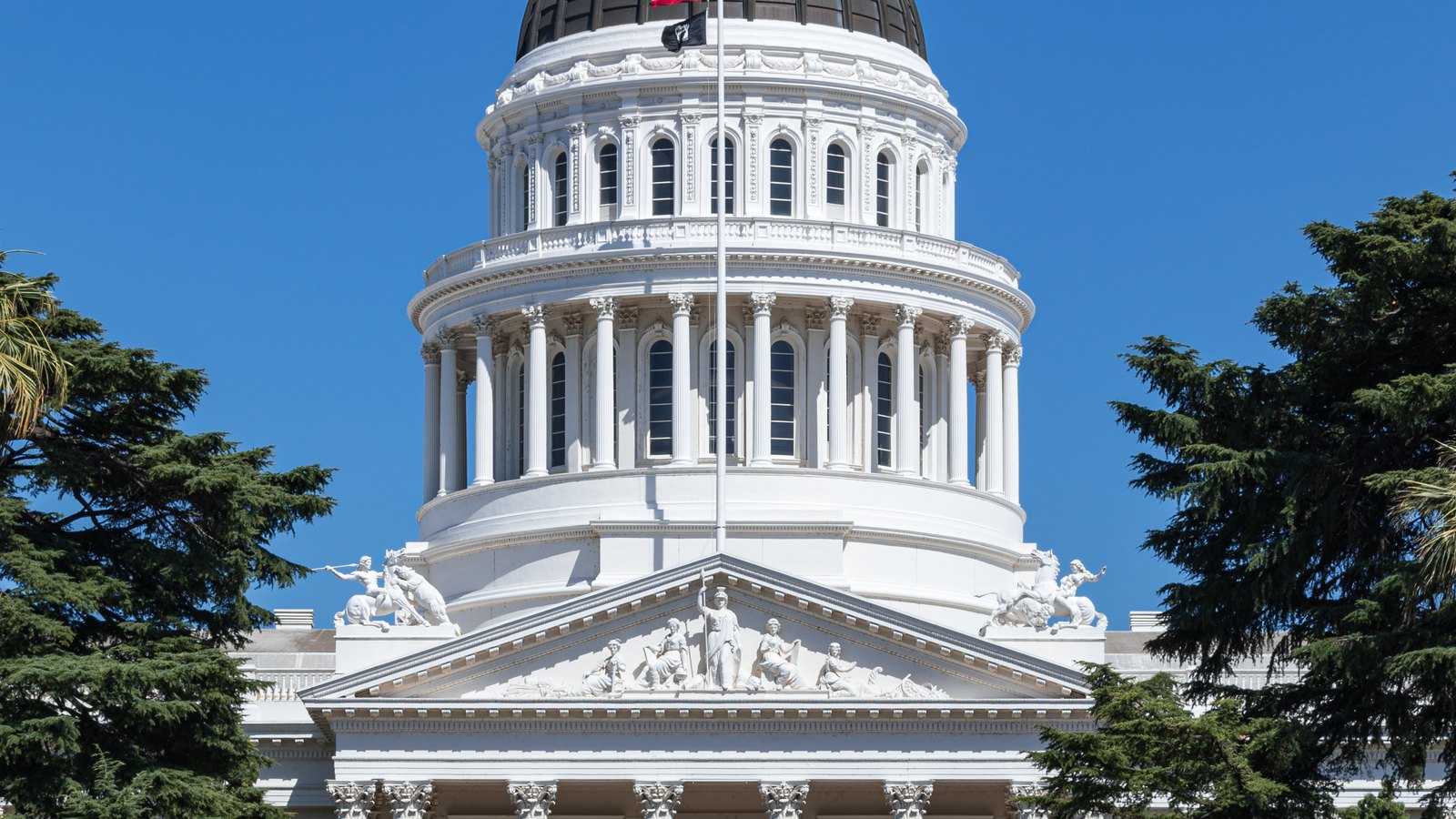 The white-columned rotunda and sculpted pediment of the California State Capitol stand against a clear blue sky.