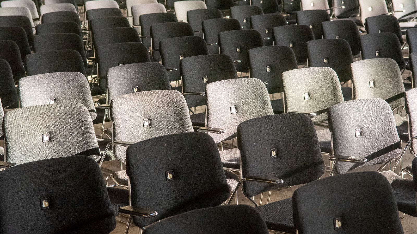 Rows of empty upholstered stacking chairs in alternating grey and black, seen at an angle across a plain indoor floor, each with a small metal writing tablet folded at the side. No person, no window, no signage and nothing identifying a place appears. A generic photograph of an empty meeting room, not a picture of Calusa Harbour or of any room in this story.