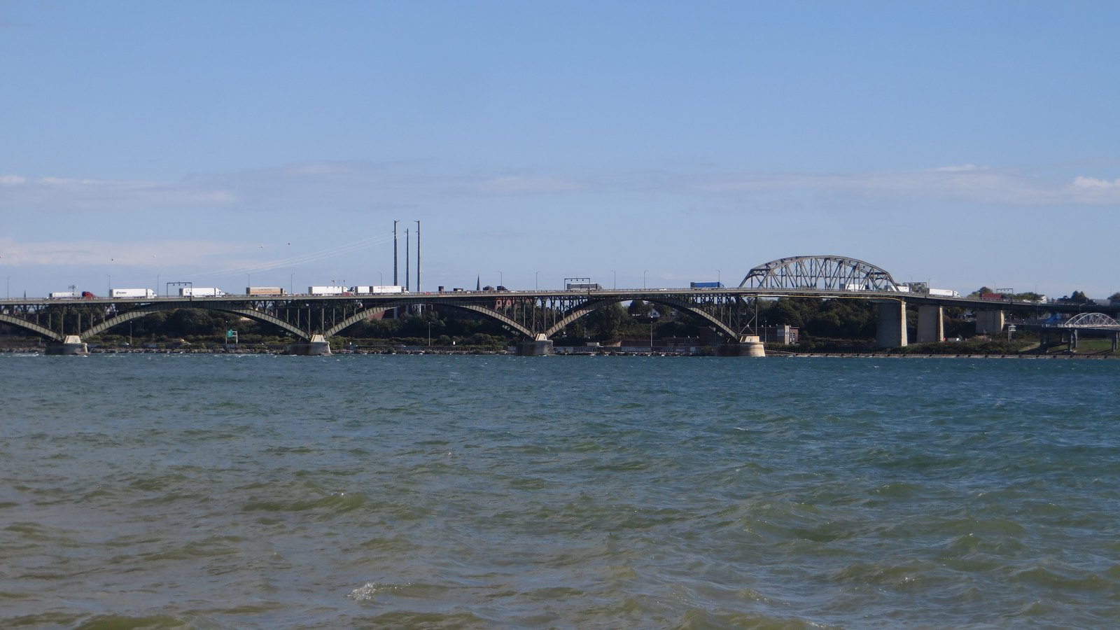 The Peace Bridge carrying a line of transport trucks and trailers across the Niagara River between Buffalo, New York and Fort Erie, Ontario, photographed from the water under a clear sky. An illustrative photograph, not taken in connection with these proclamations.