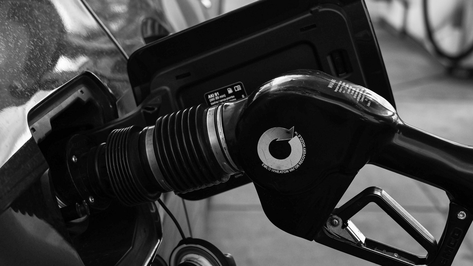 A black-and-white close-up of a fuel nozzle inserted into a car's filler neck at a pump.
