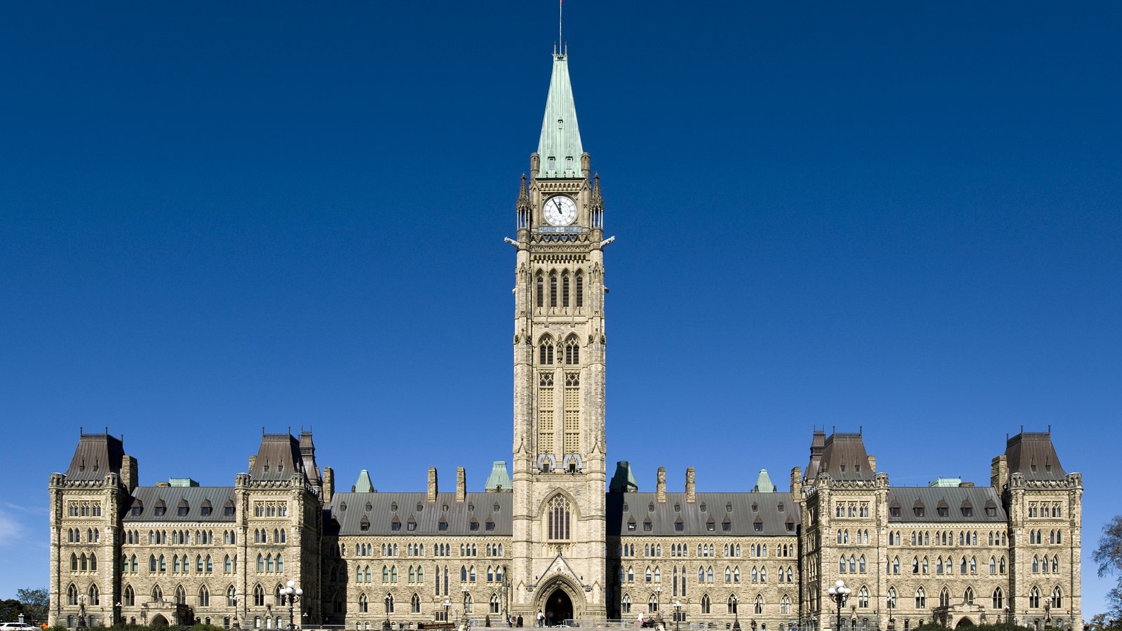 The Centre Block of the Canadian Parliament in Ottawa, photographed straight on from the lawn under a cloudless deep blue sky. The Gothic revival sandstone facade runs the full width of the frame, with steep dark roofs and pinnacled corner towers at each end, and the Peace Tower rising in the middle to a green copper spire above a white clock face. Ornamental lamp standards and a low railing line the bottom of the frame, with a few distant figures on the walkway. No signage, logo or lettering is legible anywhere in the picture.