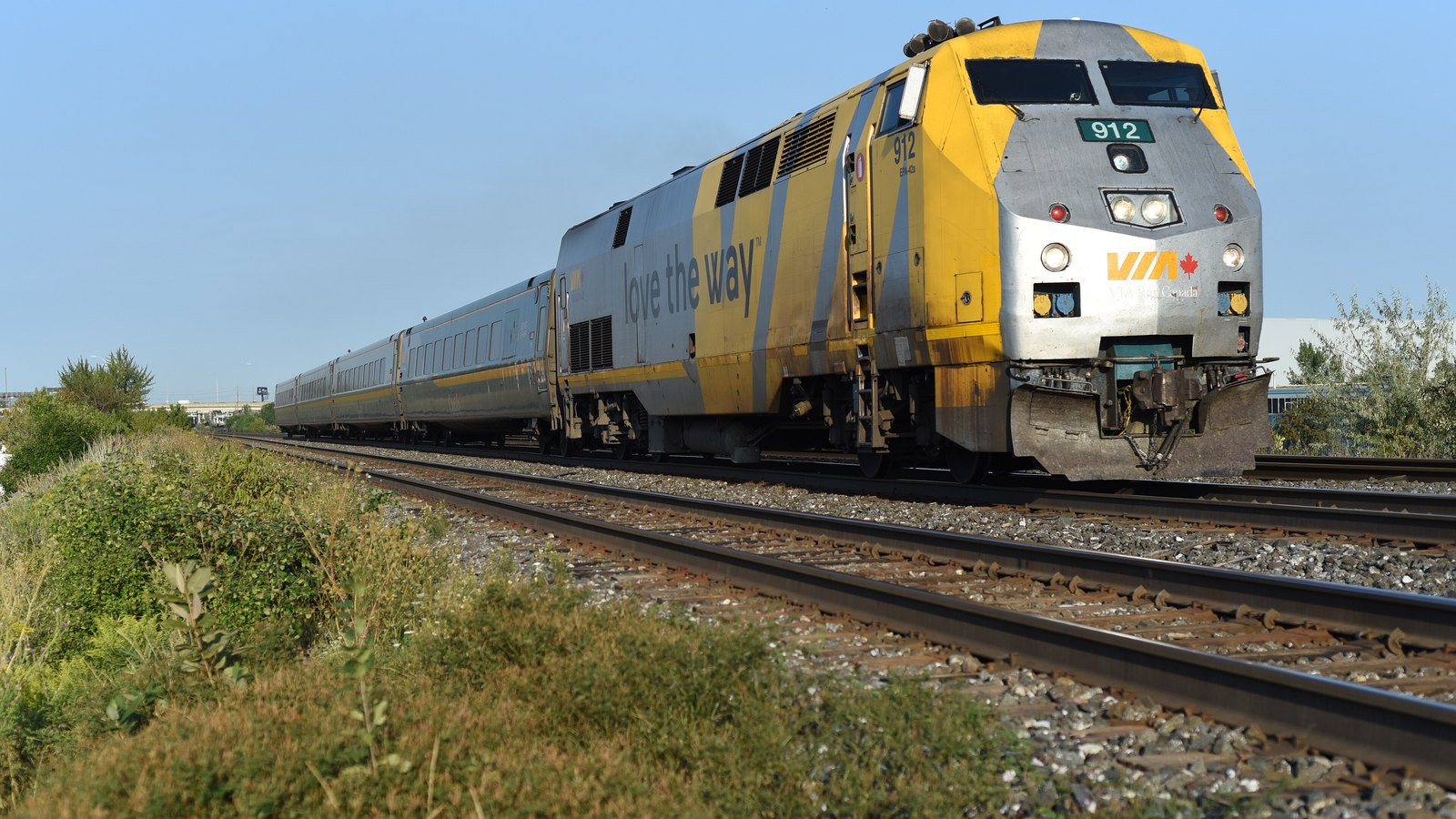 An existing VIA Rail passenger train travelling on open track near Brampton, Ontario, led by a silver and yellow locomotive under a clear sky.