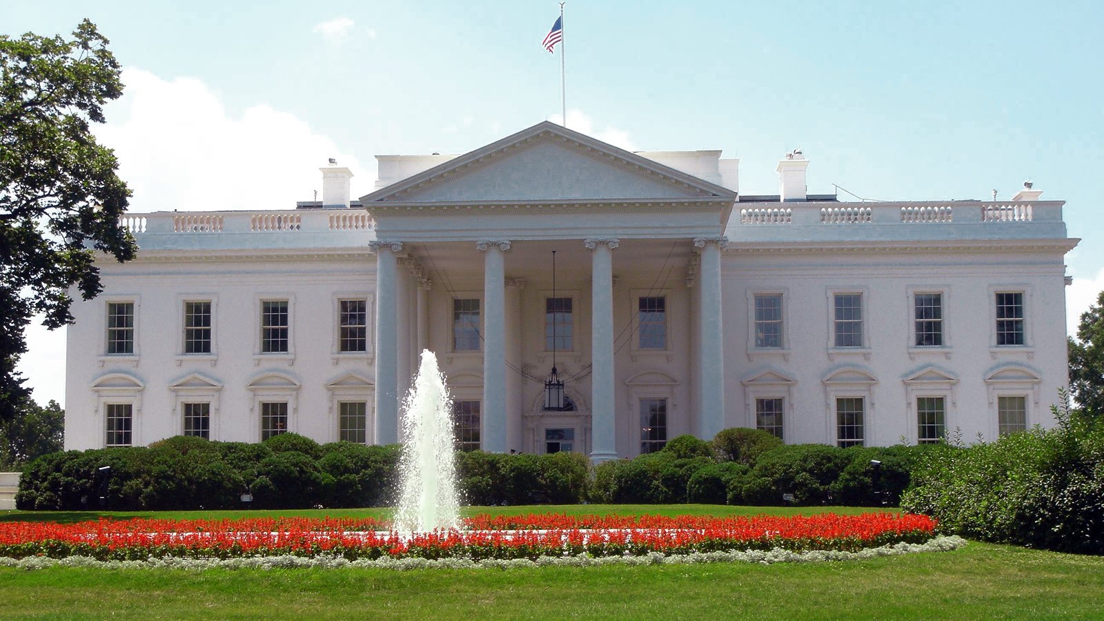 The north front of the White House stands behind a fountain and red flowerbeds beneath an American flag.