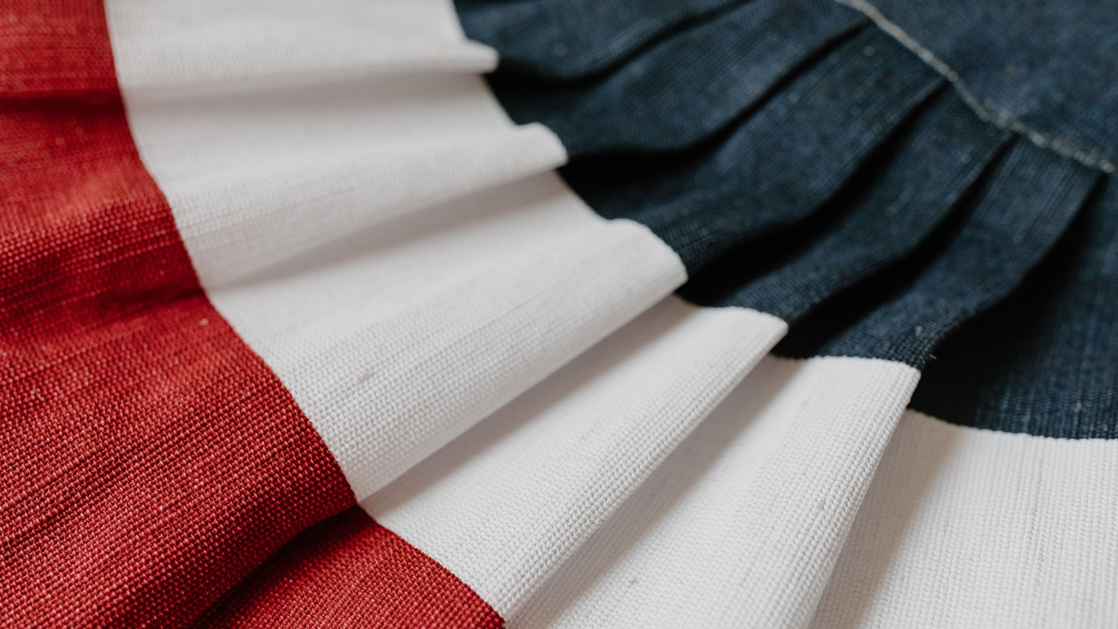 Close macro of pleated red, white and navy cotton bunting, the coarse woven texture of each fold catching soft light.