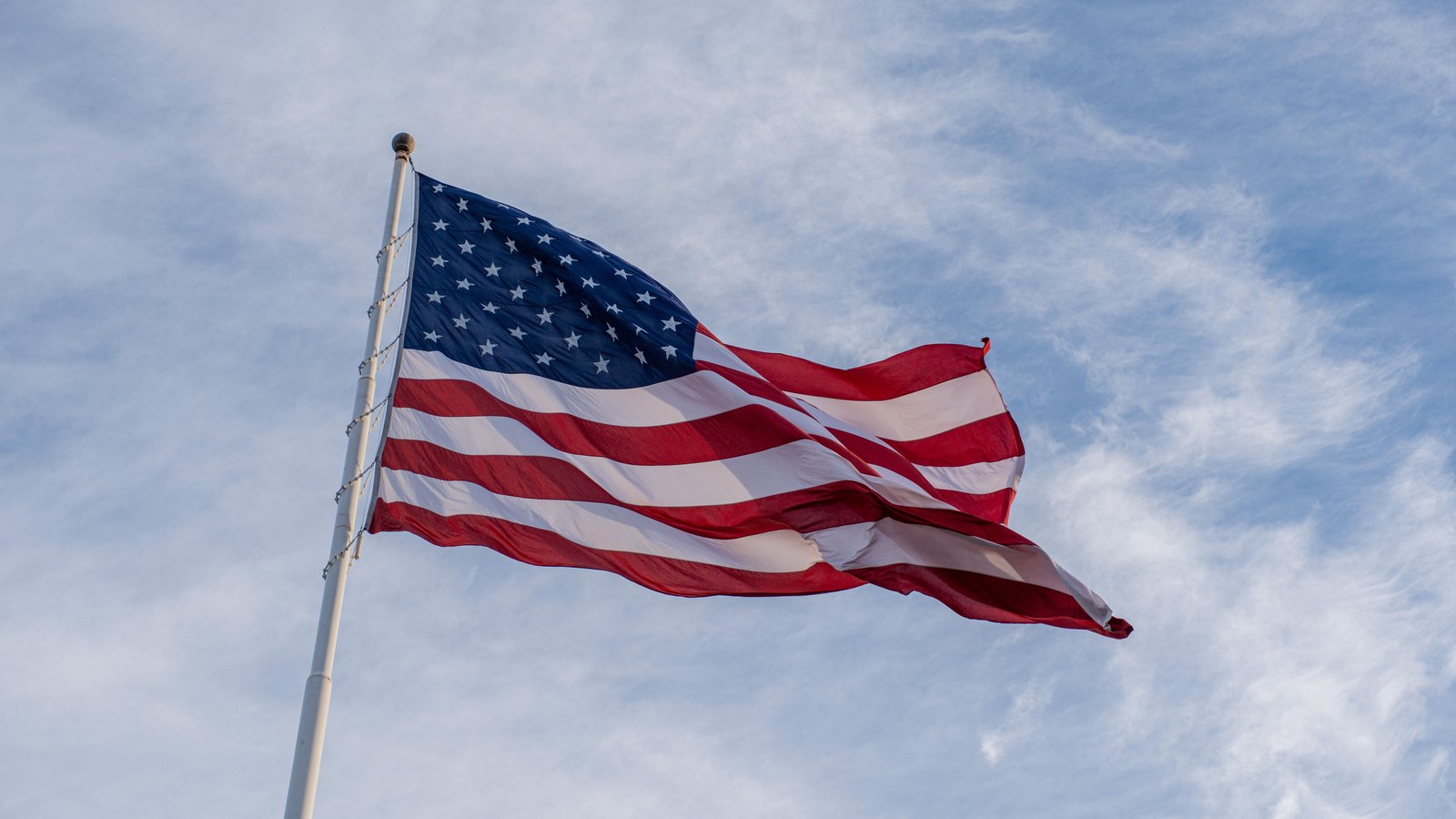 An American flag flying from a white flagpole against a pale cloudy sky, with the halyard and attachment hardware visible along the pole.