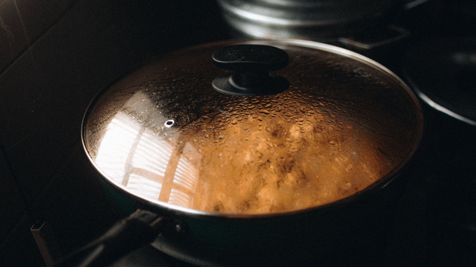 Condensation covers a glass lid over a steaming pan on a stovetop.
