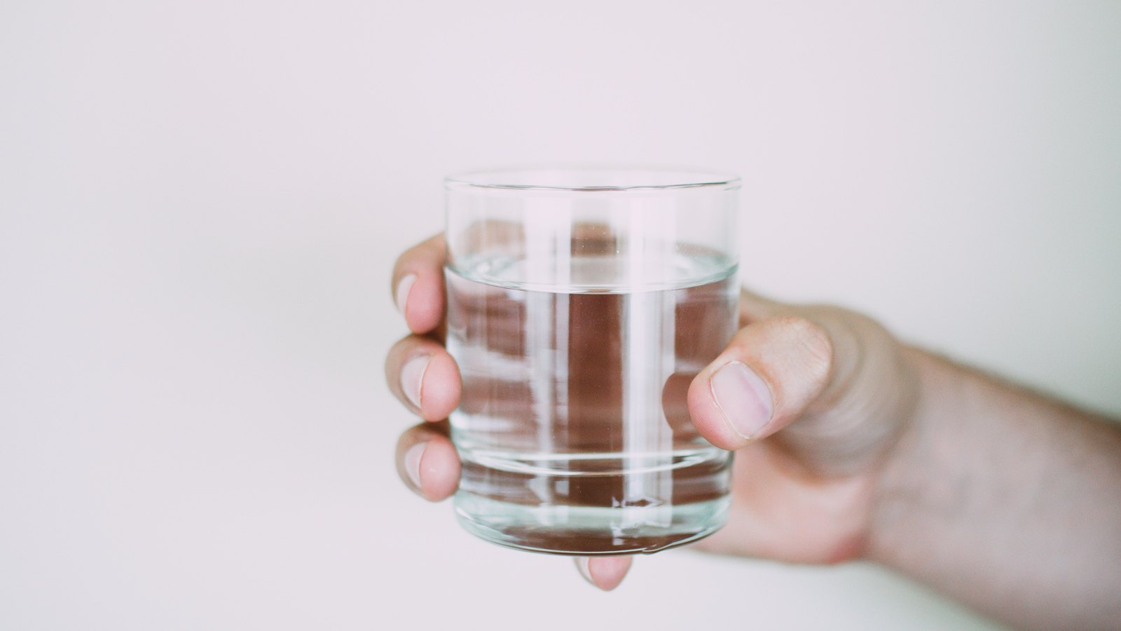 A hand holds a plain clear tumbler of water against a blank off-white wall. Only the hand and part of the forearm are in frame, so no face and no identifiable person appears, and there is no signage, brand mark, address, plate or legible text anywhere in the picture. A generic illustration of drinking water, not a photograph of this event.