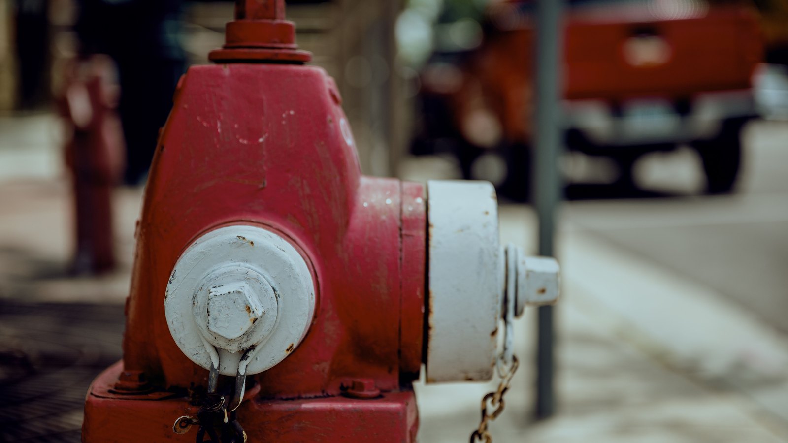 A weathered red and white fire hydrant fills the left of the frame in close focus, its cap chains hanging loose. Behind it the street is thrown well out of focus, with the red shape of a parked pickup and a pole just readable. There is no signage, brand mark, address, plate or legible text anywhere in the picture, no face and no identifiable person, and no identifiable place. A generic illustration of a drinking water story, not a photograph of this event.