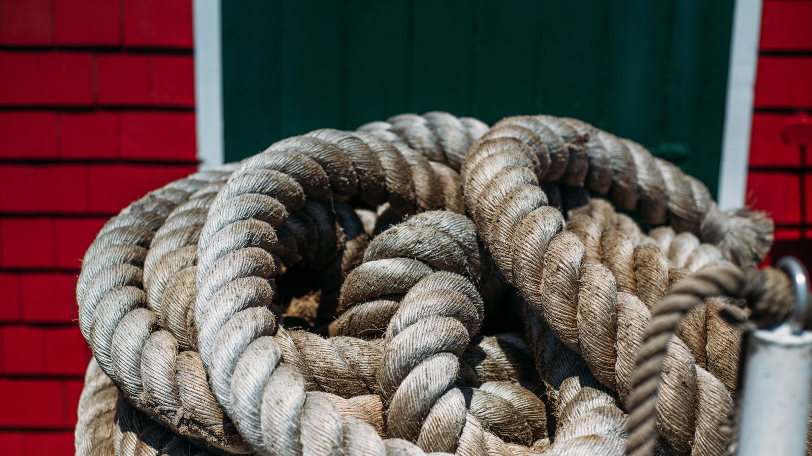 A heavy three strand mooring rope coiled in loose loops, its pale fibres worn grey, filling the frame against an out of focus red shingled wall and a dark green door.