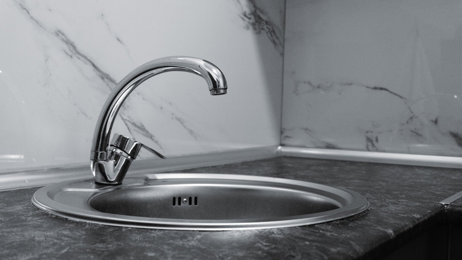 A chrome mixer tap curving over a round stainless steel kitchen sink set into a dark speckled worktop, with a plain marble patterned splashback behind it. The tap is off and the sink is empty and dry. There is no person in the frame, no signage, no lettering and no brand mark, and no place can be identified from it. A generic illustration of a household tap, not a photograph of Cape Coral or of any address that was under notice, and not evidence about the condition of anyone's water.