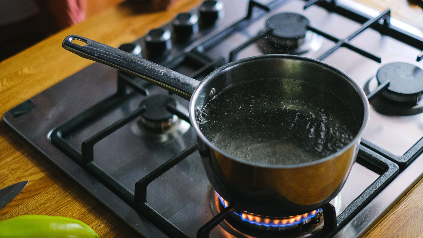A stainless steel saucepan of water at a rolling boil sits on a lit gas burner in a home kitchen, with a green pepper on the wooden worktop beside it.