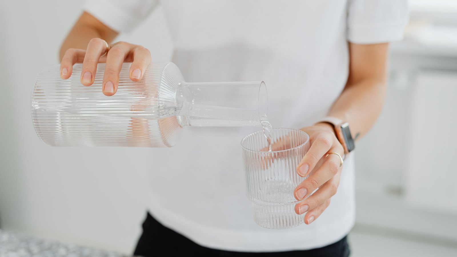 A photograph of a person in a white shirt, framed from the neck down, tipping a ribbed glass carafe of clear water into a matching ribbed tumbler held in the other hand, against a pale out of focus kitchen wall. No face, signage, lettering, brand marks or identifiable place are visible and the water is clear and colourless. A generic household illustration, not a photograph of Cape Coral or of any address under notice, and not evidence about the condition of anyone's water.