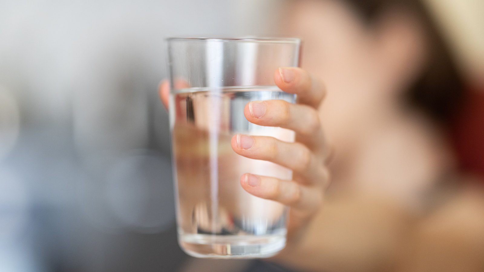 A hand holds a clear glass of water in front of a softly blurred face, not a photograph of this event.