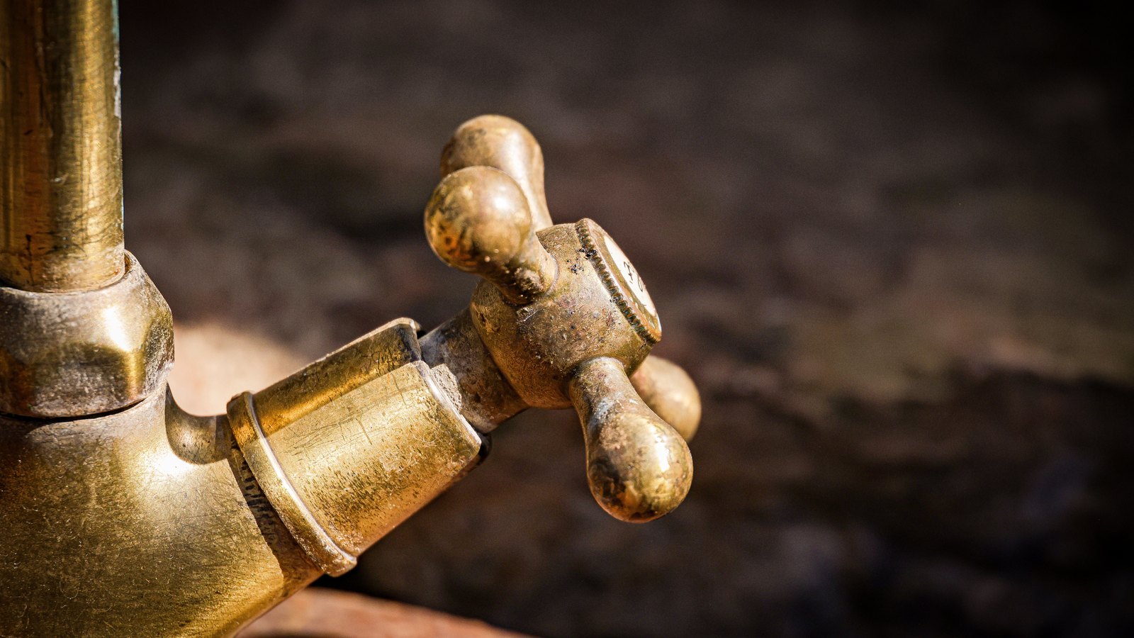 A close up photograph of an aged brass tap with two rounded cross handles, mounted on a brass pipe elbow against a dark blurred background. The tap is closed and dry, with no water running from it. There is no text, signage, brand mark, person, hand, food or identifiable place in the frame. A generic illustration of plumbing hardware, not a photograph of Cape Coral, of the Diplomat Parkway East work, of anyone's tap, or of anyone's water. Its weakness is stated rather than glossed: the fixture is an old outdoor style brass tap rather than anything typical of a Southwest Florida home, and the brass is tarnished with age, which should not be read as a statement about the condition of anyone's water supply.