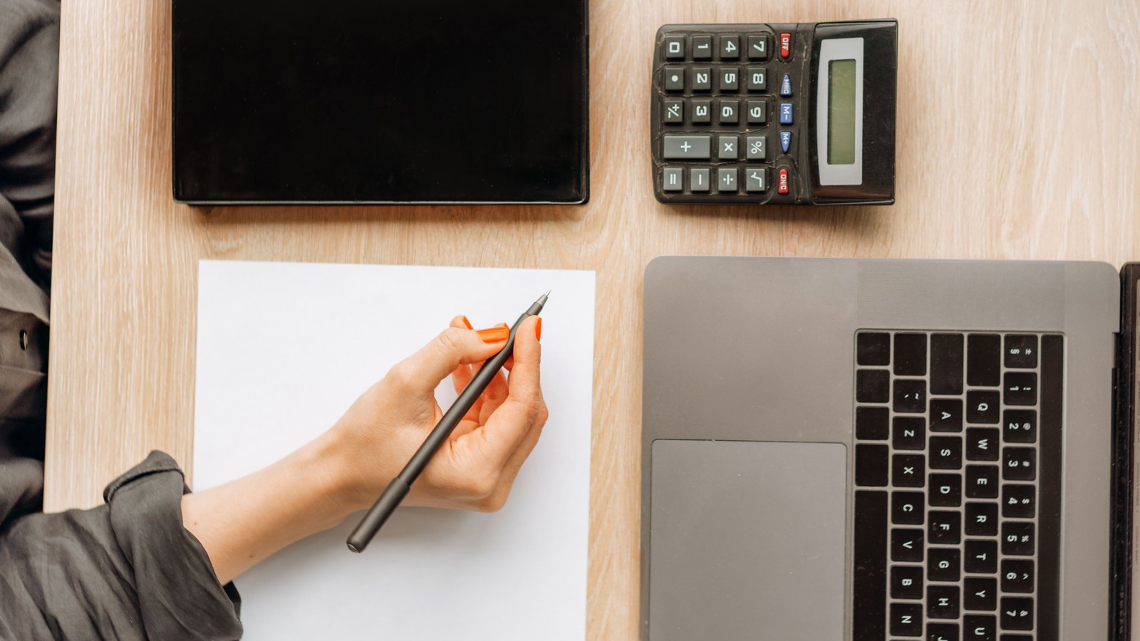 A desk seen from above, with a hand holding a pen over a blank sheet of paper beside a calculator and a laptop.