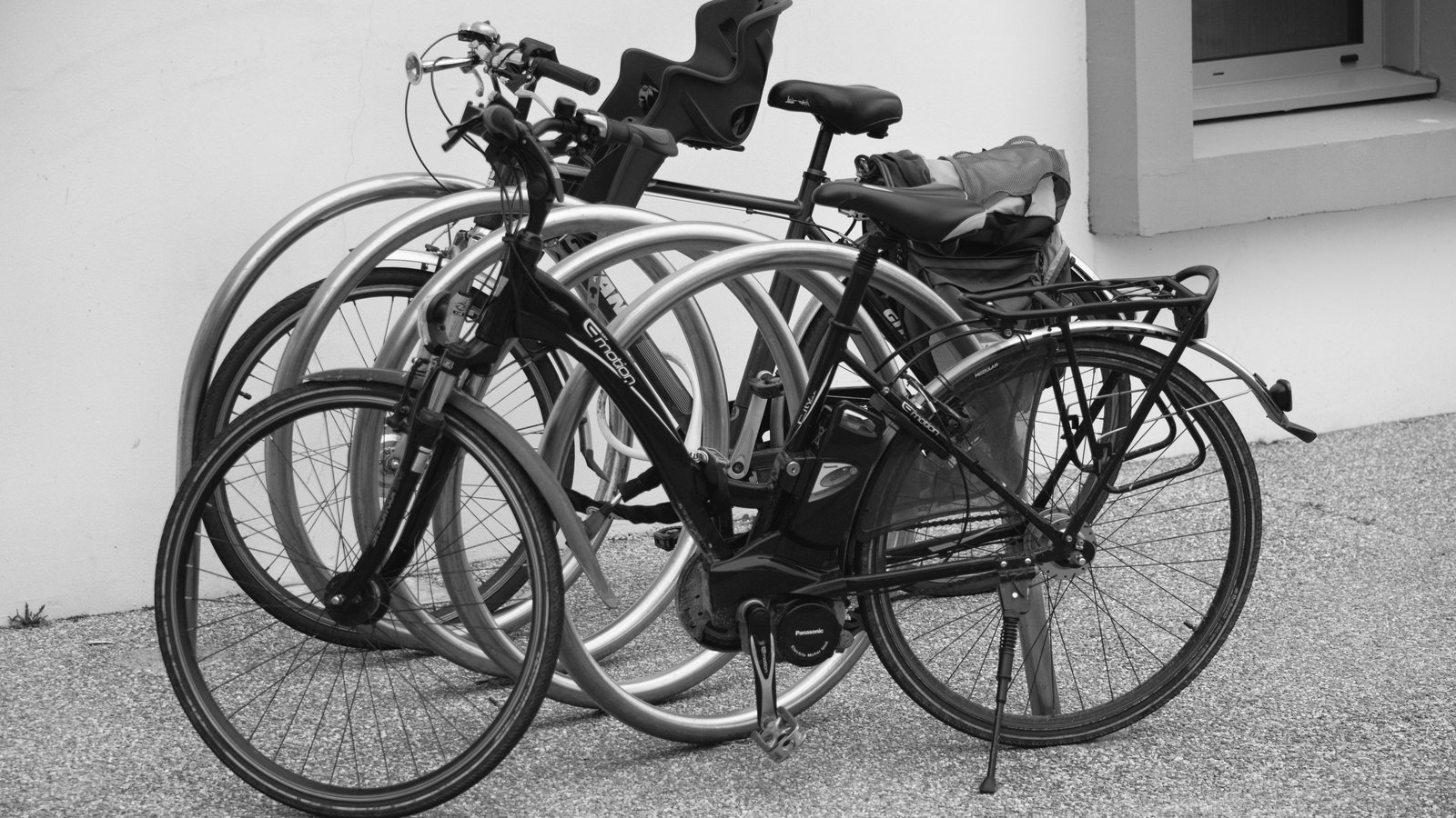A black and white photograph of two electric bicycles parked in a metal cycle rack against a plain rendered wall, the nearer one fitted with a mid-drive motor, a rear luggage rack and a soft bag strapped over the saddle. There is nobody in the picture and no street name, house number, number plate, signage or agency marking anywhere in the frame. Stock photograph, not a photograph of this event.