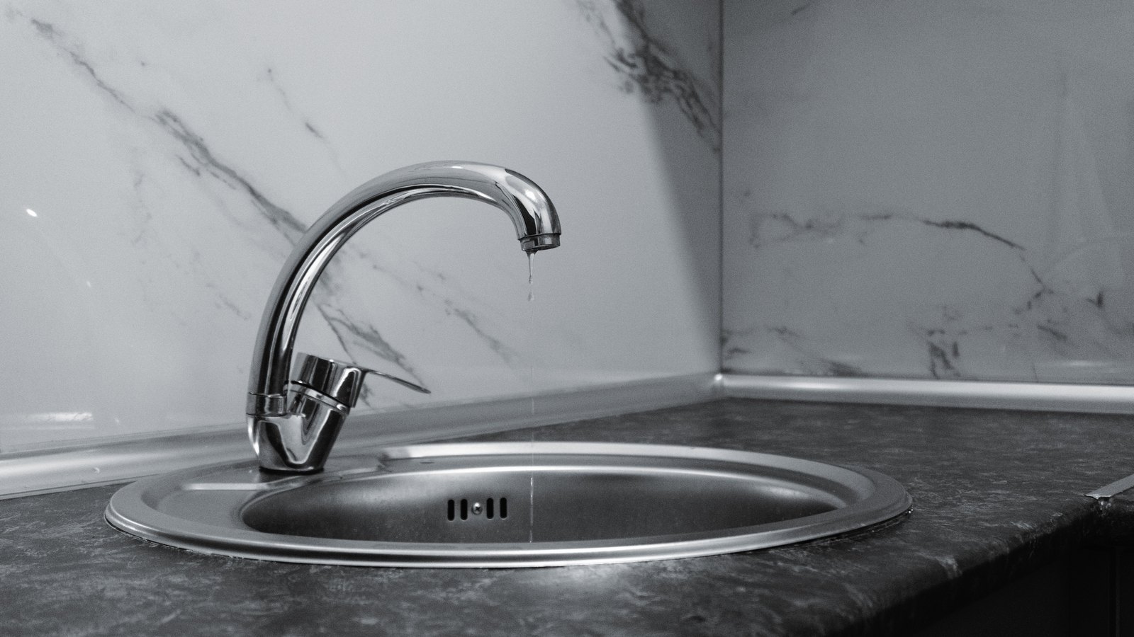 A black and white photograph of a chrome kitchen mixer tap dripping into a round stainless steel sink set in a dark stone counter, with a marble backsplash behind it, no people, signage or text in view. A generic illustration, not a photograph of Cape Coral.