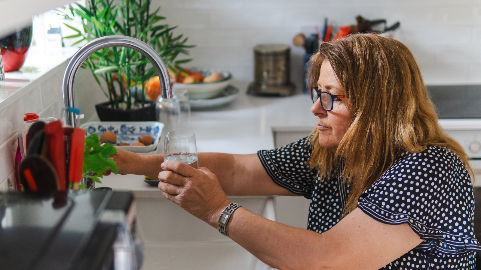 A woman in a navy polka dot blouse and dark rimmed glasses holds a clear tumbler under a running chrome kitchen tap, filling it with water. Behind her are a white worktop, a potted plant, a fruit bowl and a dish of eggs. No signage, place name or legible text is visible anywhere in the frame and no location can be identified from it. Pexels titles it a woman filling a glass with water in a home kitchen. A generic stock illustration of a tap being used for drinking water, not a photograph of Cape Coral, of Fairmont Cove Court or of any resident affected by this notice.