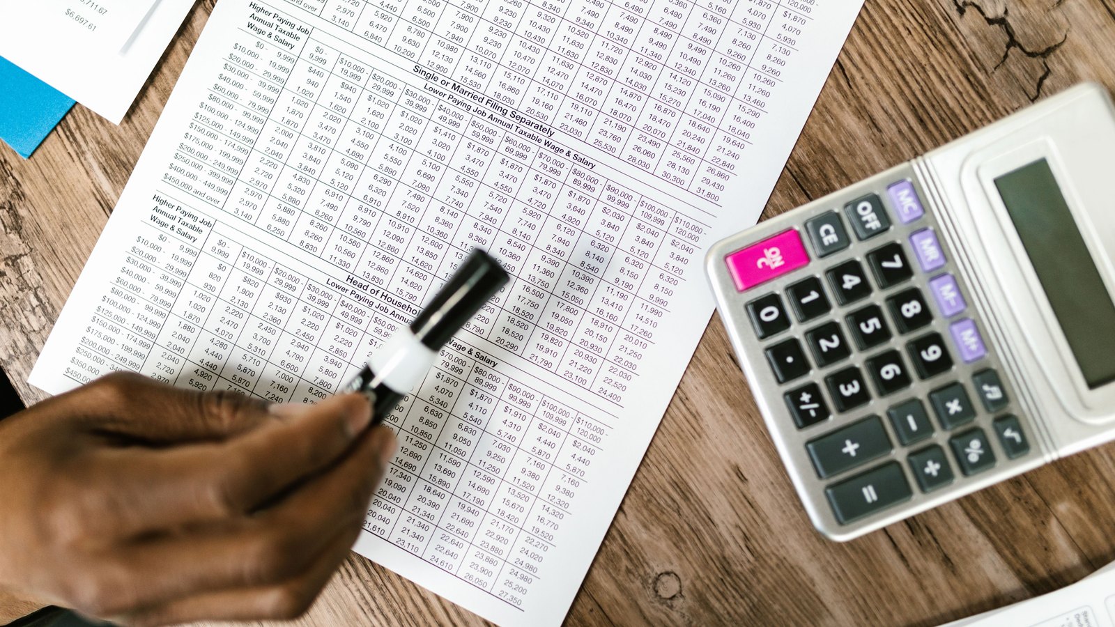 Overhead view of a printed sheet of tax rate tables on a wooden desk beside a white pocket calculator, with one hand holding a marker over the columns of figures. No face, no logo, no place and no named party are visible in the frame. Illustrative only, not a photograph of this event.