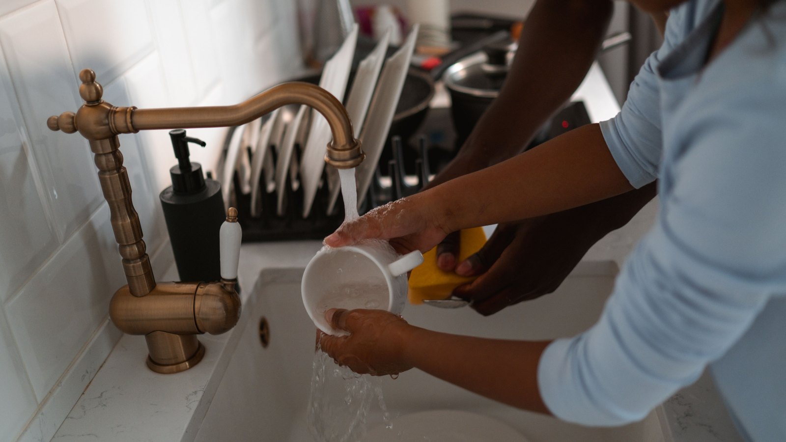 Water running from a brass kitchen tap over a white mug held under the stream, with a soap dispenser and a rack of drying plates behind it. Two pairs of hands and forearms are in frame and nothing above the elbow, so no face and no identifiable person appears, and there is no signage, no brand mark, no address and no legible text anywhere in the picture. A generic illustration of tap water and washing up, not a photograph of any address under a notice.