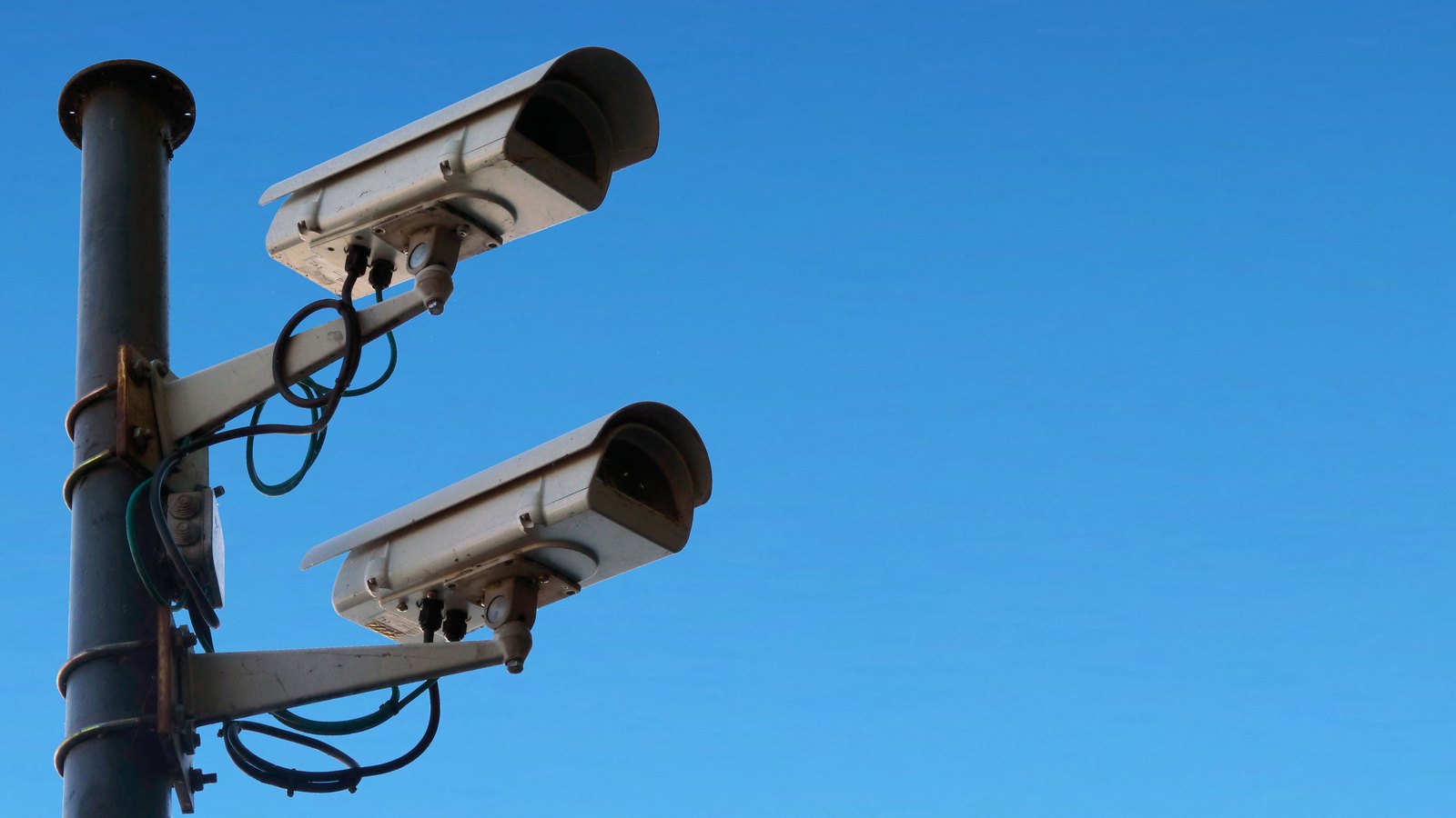 Two white box shaped surveillance cameras on separate brackets clamped to a dark grey metal pole, photographed from below against a plain clear blue sky with loose cable loops hanging beneath each mounting, and no people, vehicles, buildings, signs, brand marks or text in view. A generic illustration, not a photograph of the Cape Coral cameras.