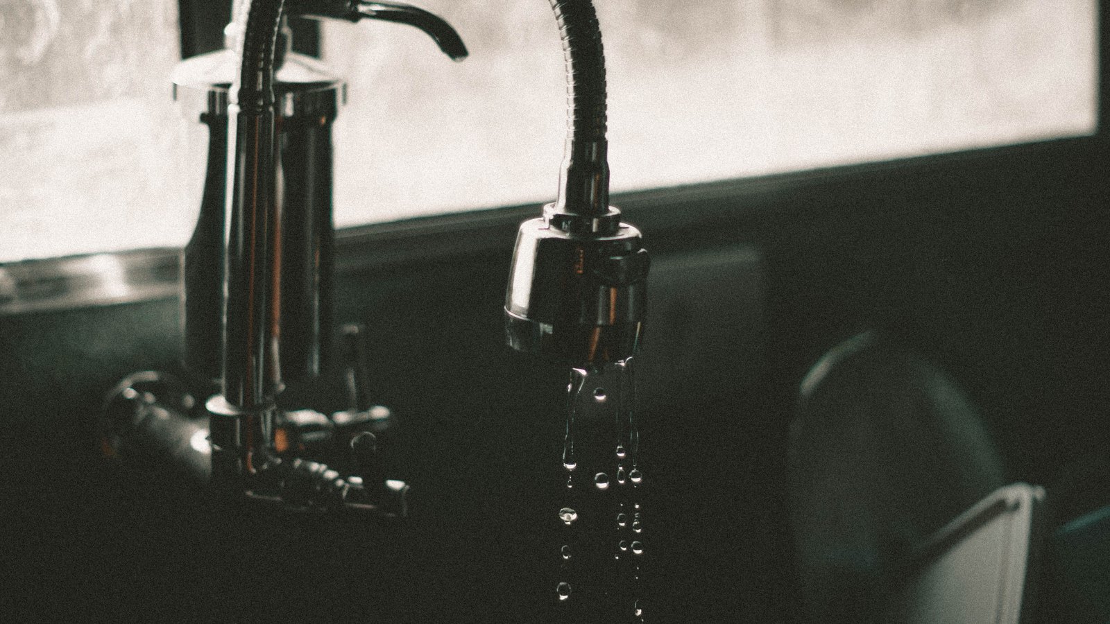 A photograph of a chrome pull down kitchen faucet in low light, its spout releasing a thin broken stream of water into a dark sink, with a bright window behind it out of focus. No people, signage, lettering or brand marks are visible and no place can be identified from it. A generic household illustration, not a photograph of Cape Coral or of any address under notice, and not evidence about the condition of anyone's water.
