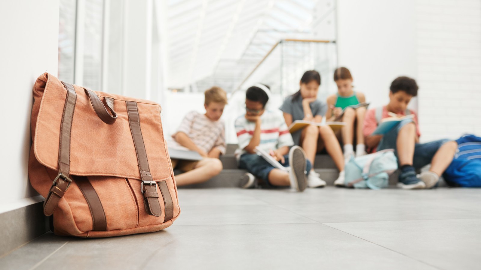 An orange canvas school bag propped against a white wall in the foreground of a bright indoor corridor, with several out of focus children sitting on steps reading in the far background. No signage, no street and nothing identifying a place appears. A generic back to school photograph, not a picture of anyone or anywhere in this story.
