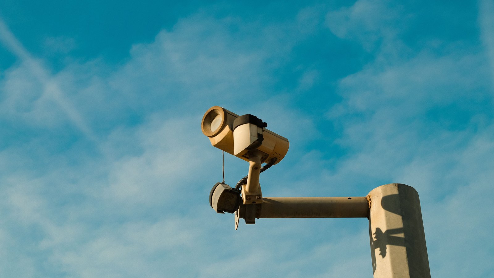 A pale metal surveillance camera and a smaller sensor unit mounted on a horizontal arm extending from a concrete pole, photographed from below against a blue sky with thin high cloud, with no people, vehicles, buildings, signs or text in view.